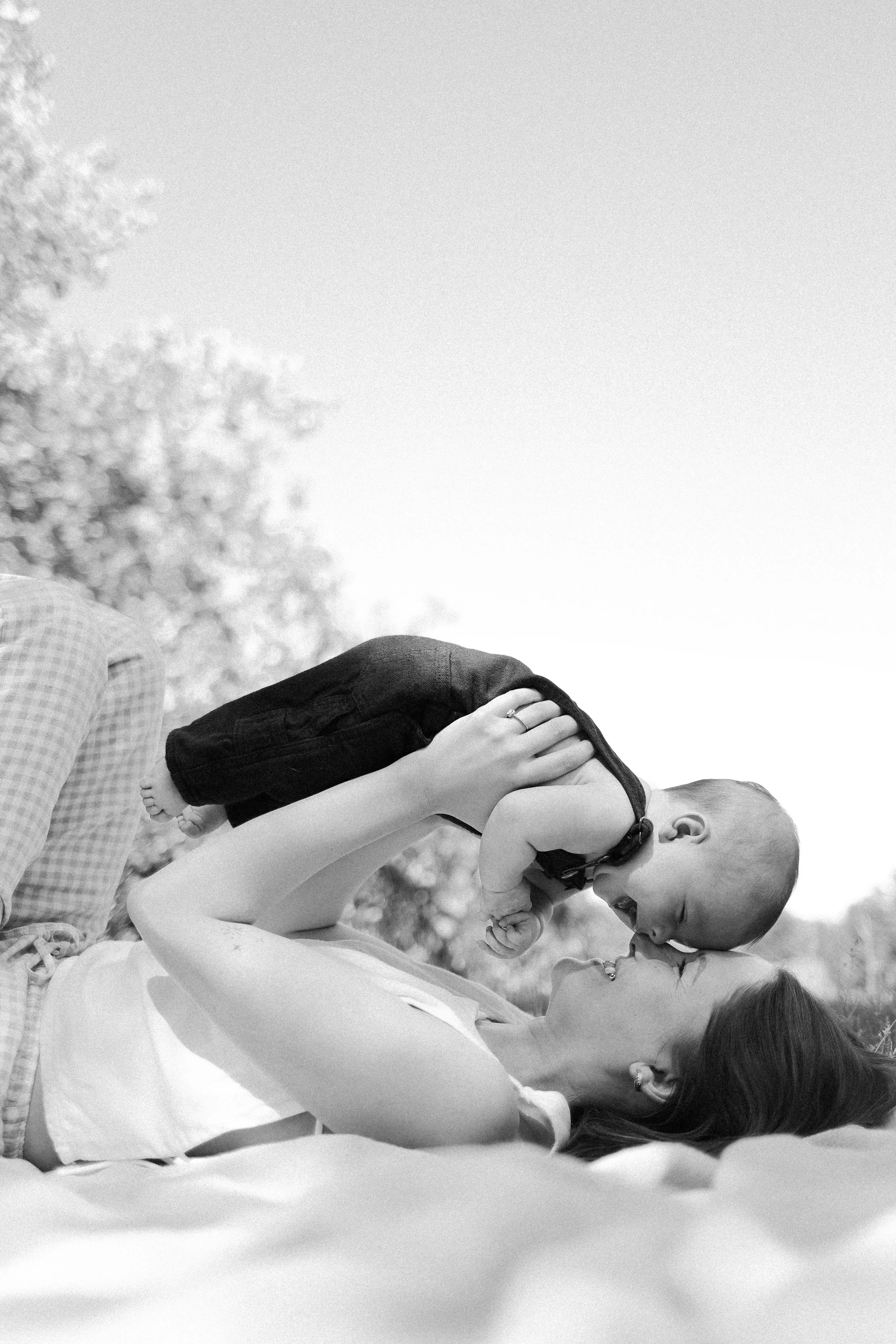 A young woman with long hair is lying down outdoors holding a small child on her chest. The child is leaning in to kiss the woman on the lips. The background is blurred with trees and a person partially visible on the left side. The photo is in black