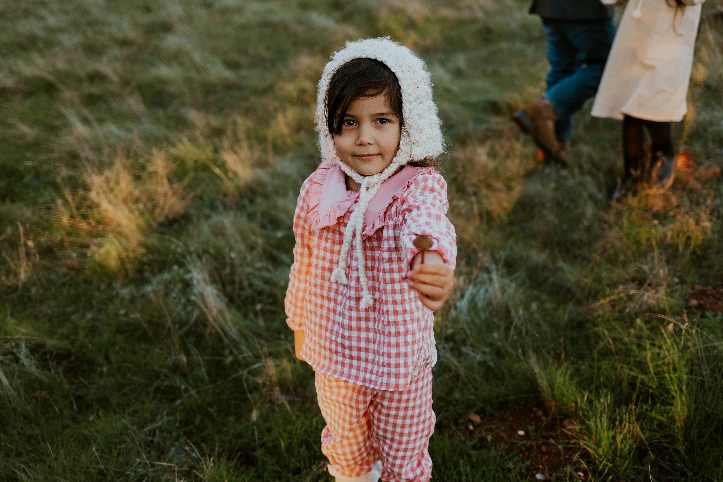 A young girl with dark hair and brown eyes, wearing a pink checkered coat and a white knit hat, standing outdoors in a grassy area.