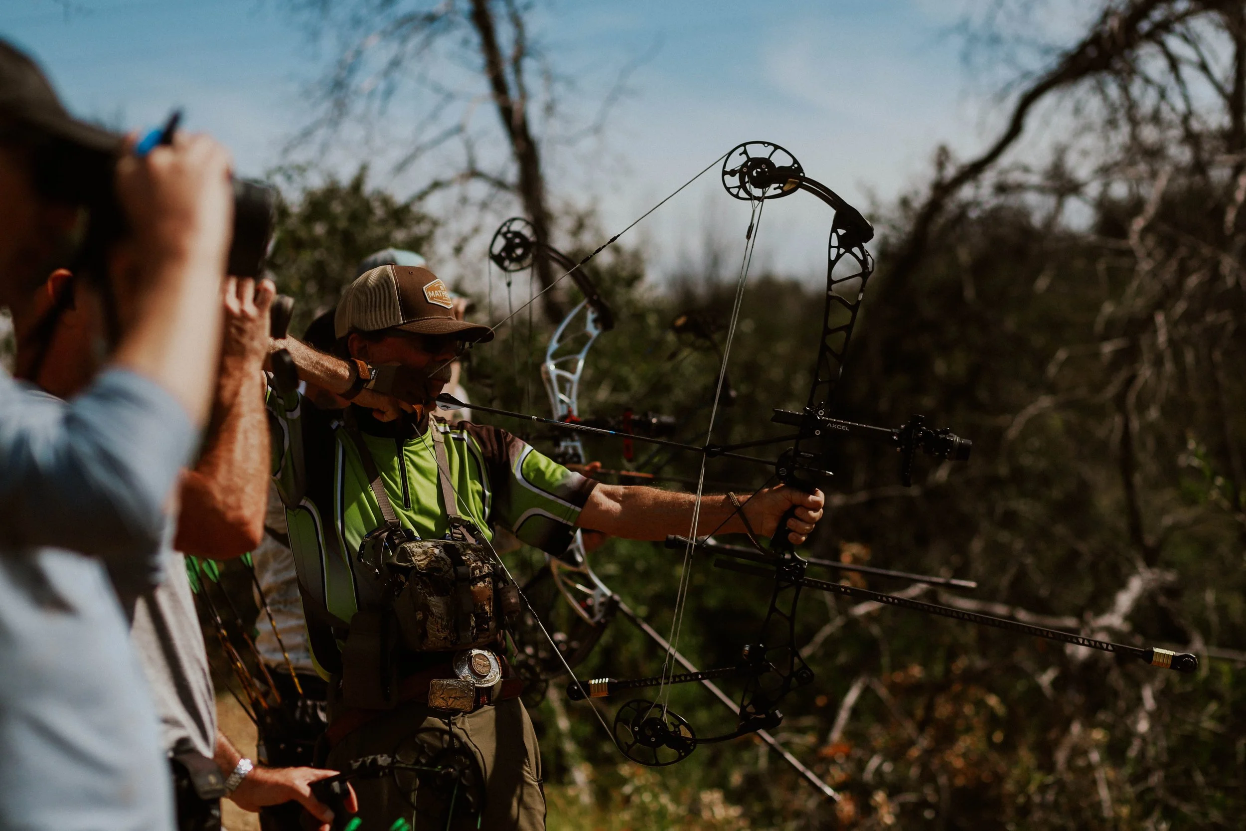 A group of people outdoors practicing archery, focusing on a man with a bow aiming at a target, with trees and a clear blue sky in the background.