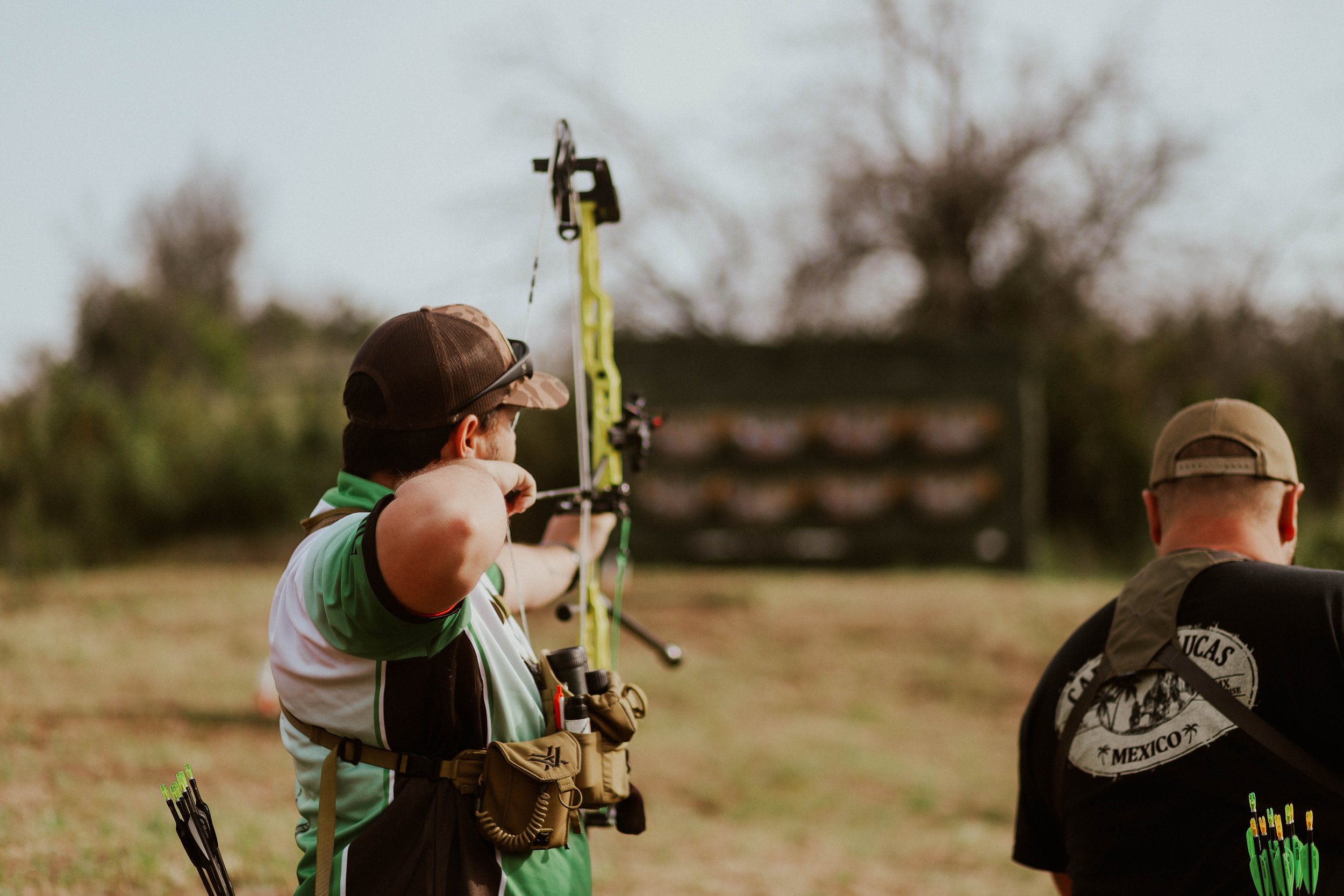 A man wearing a baseball cap and a green and white shirt is aiming a bow and arrow at an outdoor archery range. Other people are present, one wearing a black shirt with a circular logo, and trees are visible in the background.
