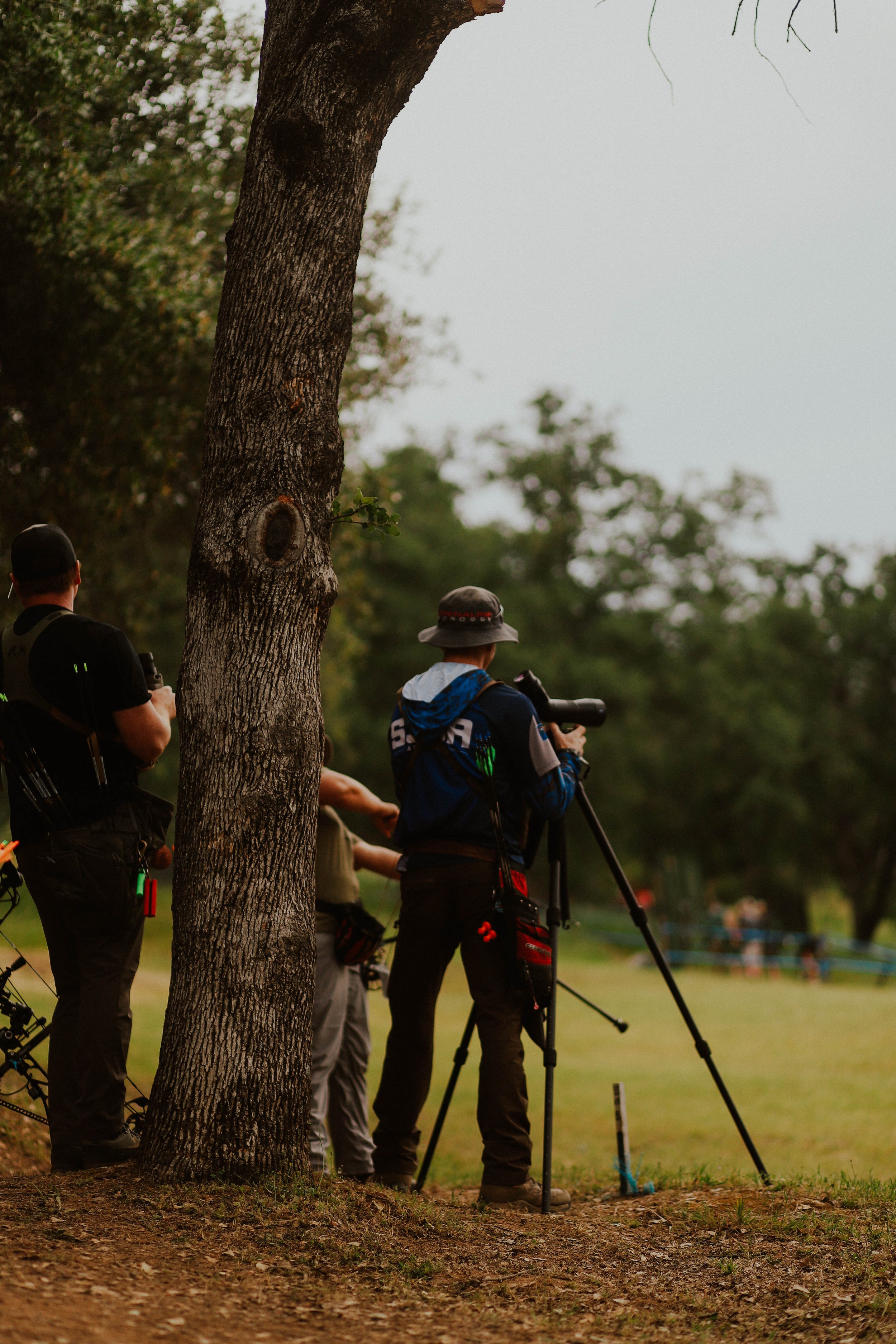 Group of people with photography equipment gathered outdoors behind a large tree, with trees and a park in the background.
