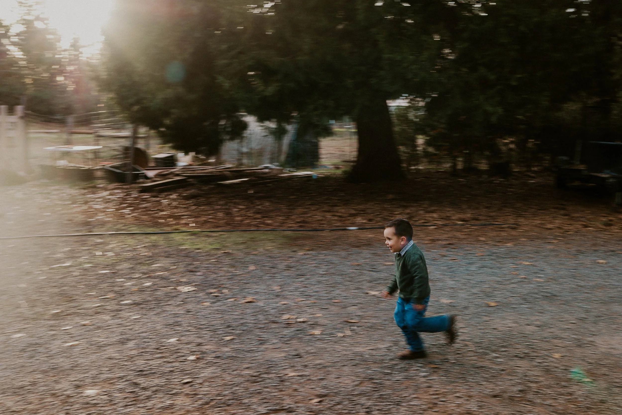 Young boy running with a soccer ball in an outdoor park with trees in the background, sun shining through the trees.