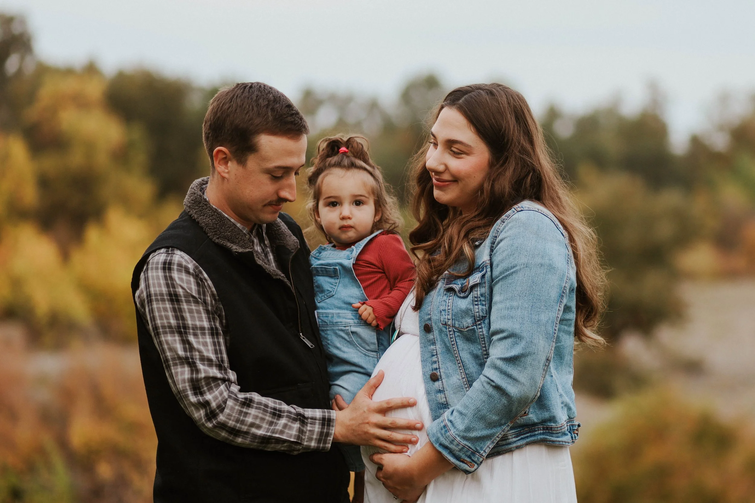 A pregnant woman and a man stand outdoors in a park with trees, with the woman holding her belly. The woman has long brown hair and is smiling, wearing a denim jacket. The man has short hair, a mustache, and is looking down at her belly, wearing a pl