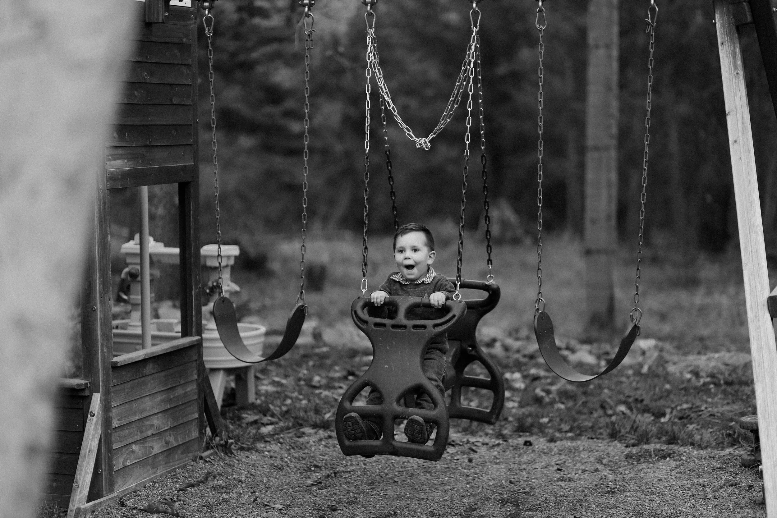 A young boy on a swing set in a playground, with trees in the background, in black and white.