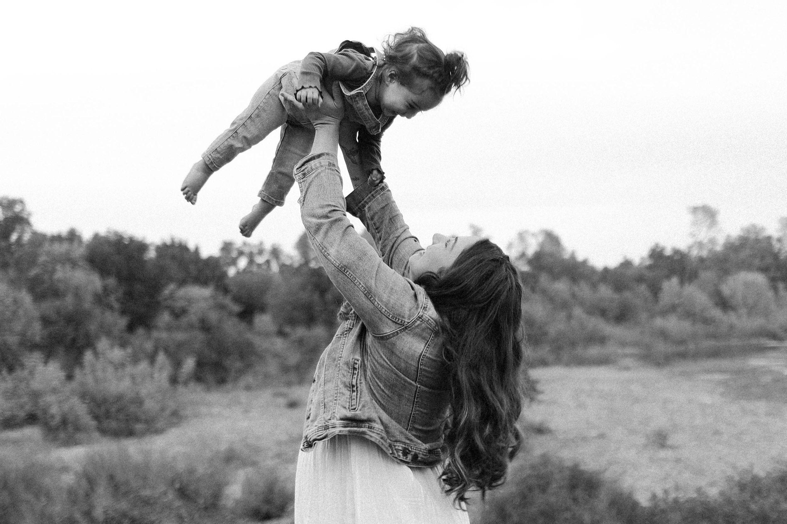 A woman holding a child in her arms outdoors, with trees and open sky in the background.