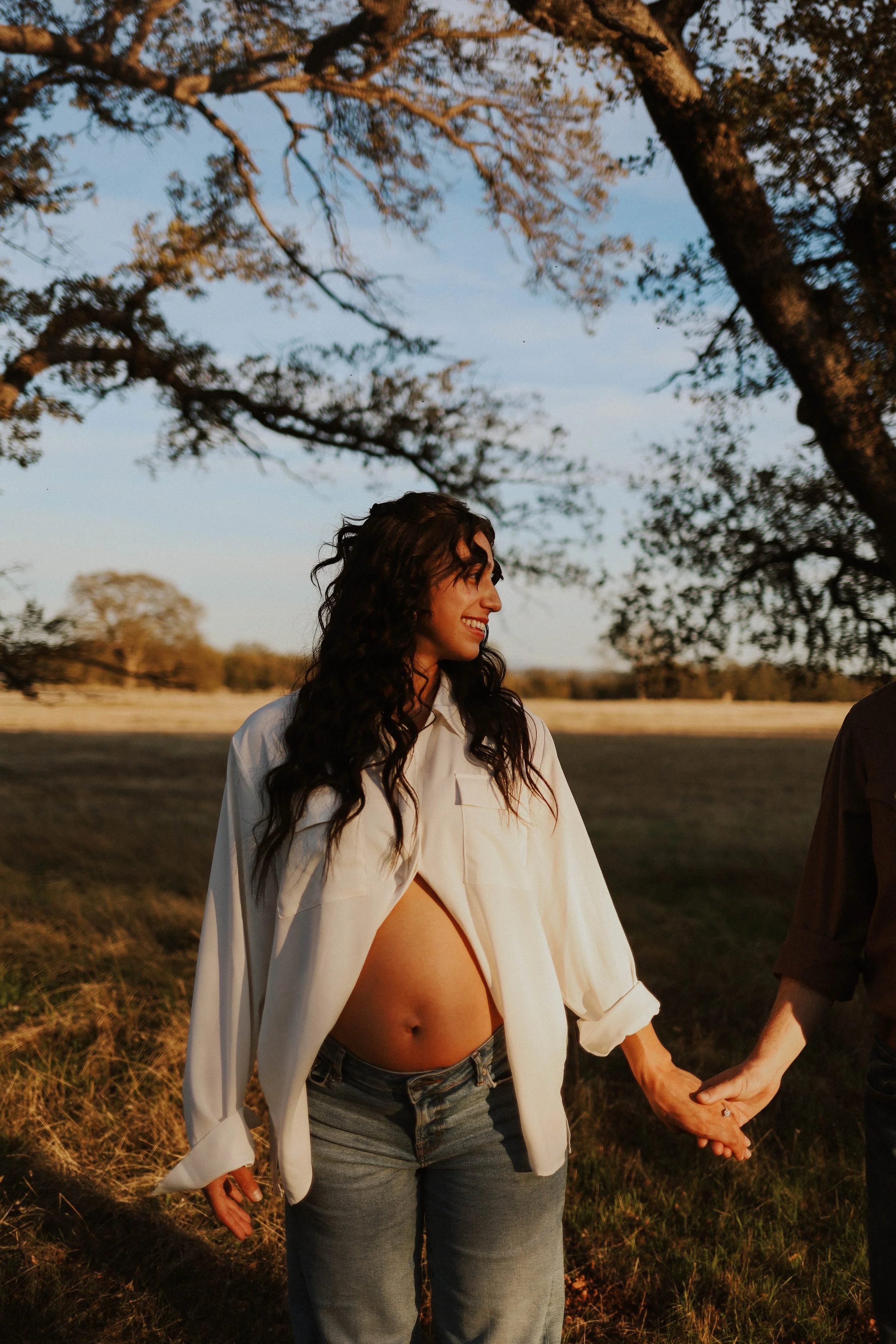 A woman with long curly hair smiling and holding hands with someone outside in a field during sunset, trees in the background.