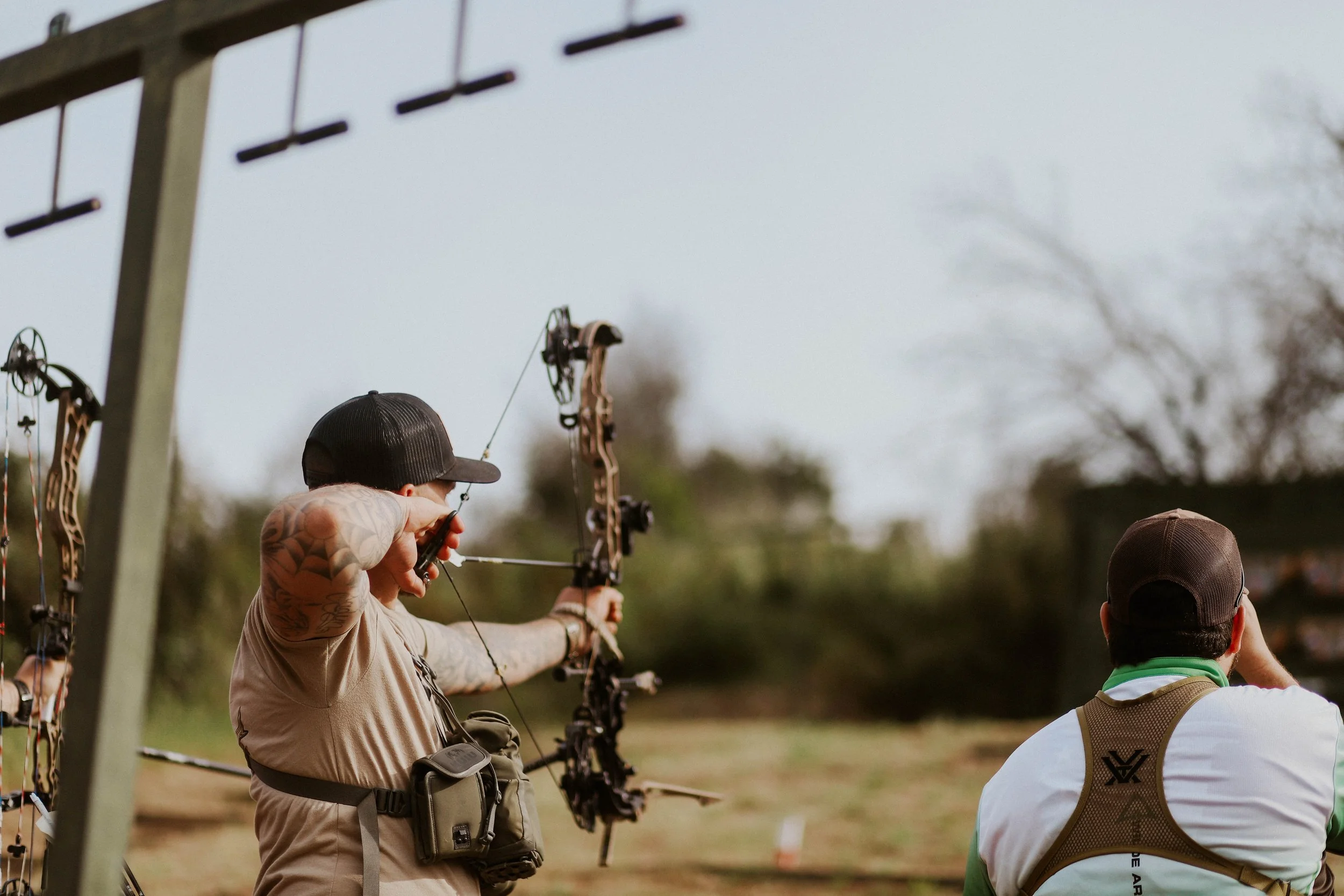 Multiple people practicing archery outdoors, drawing bows aimed at targets, with trees and a cloudy sky in the background.