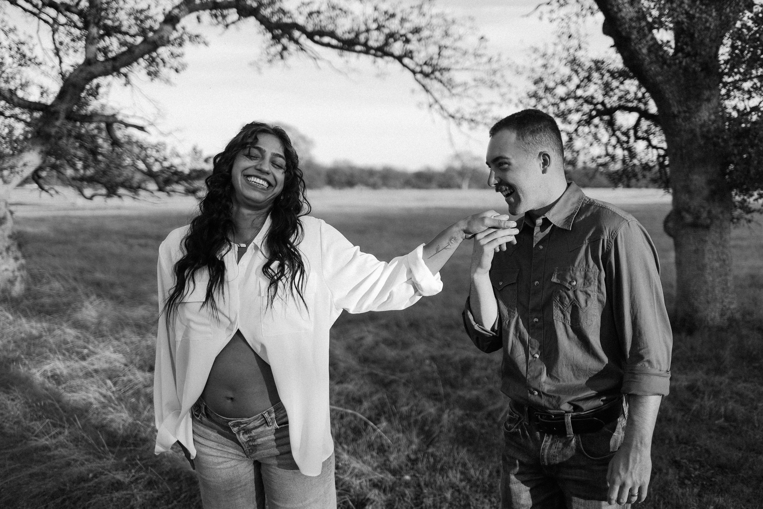 A black and white photo of a pregnant woman smiling with her eyes closed and hand in a man's hand, in an outdoor natural setting with trees in the background.