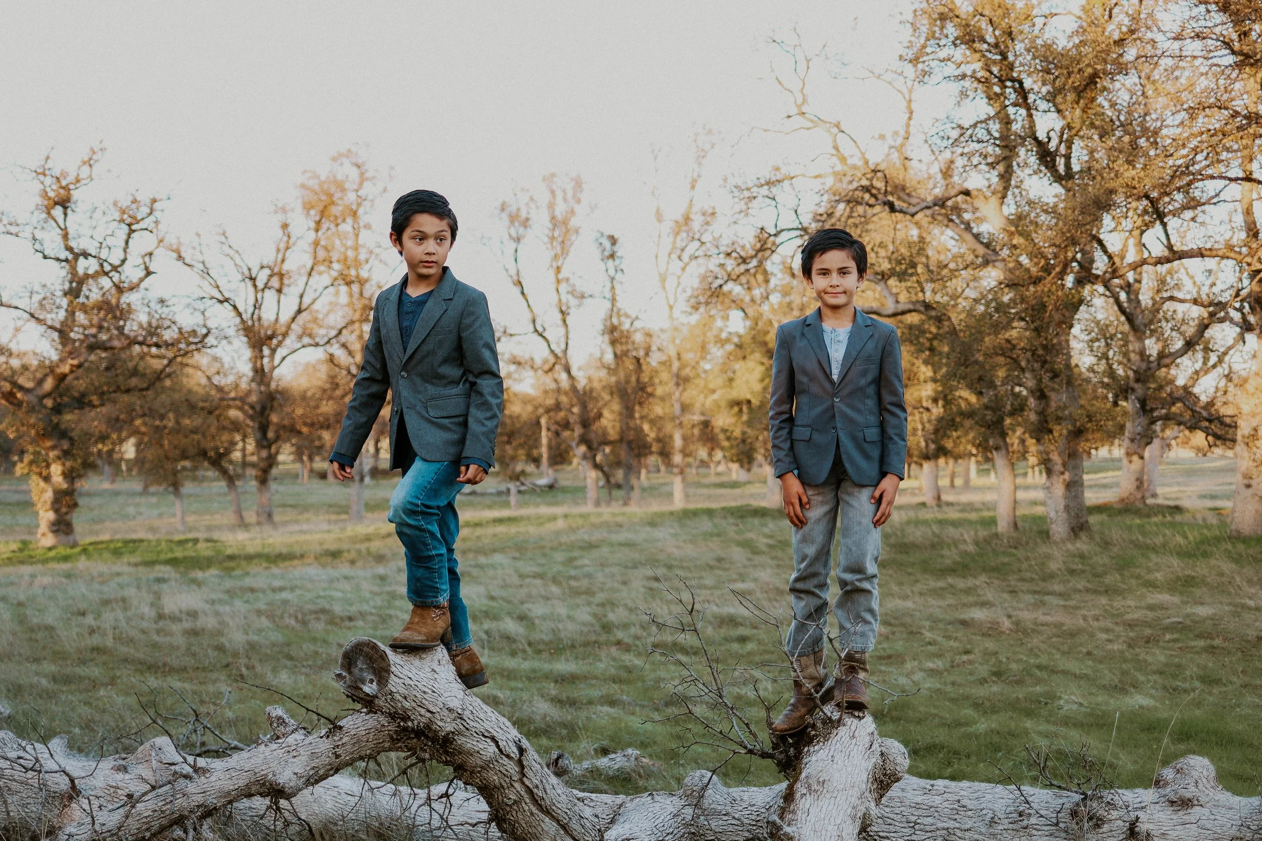 Two young boys in suits, one standing on a fallen tree trunk and the other standing on the ground, in a park with autumn-colored trees in the background.
