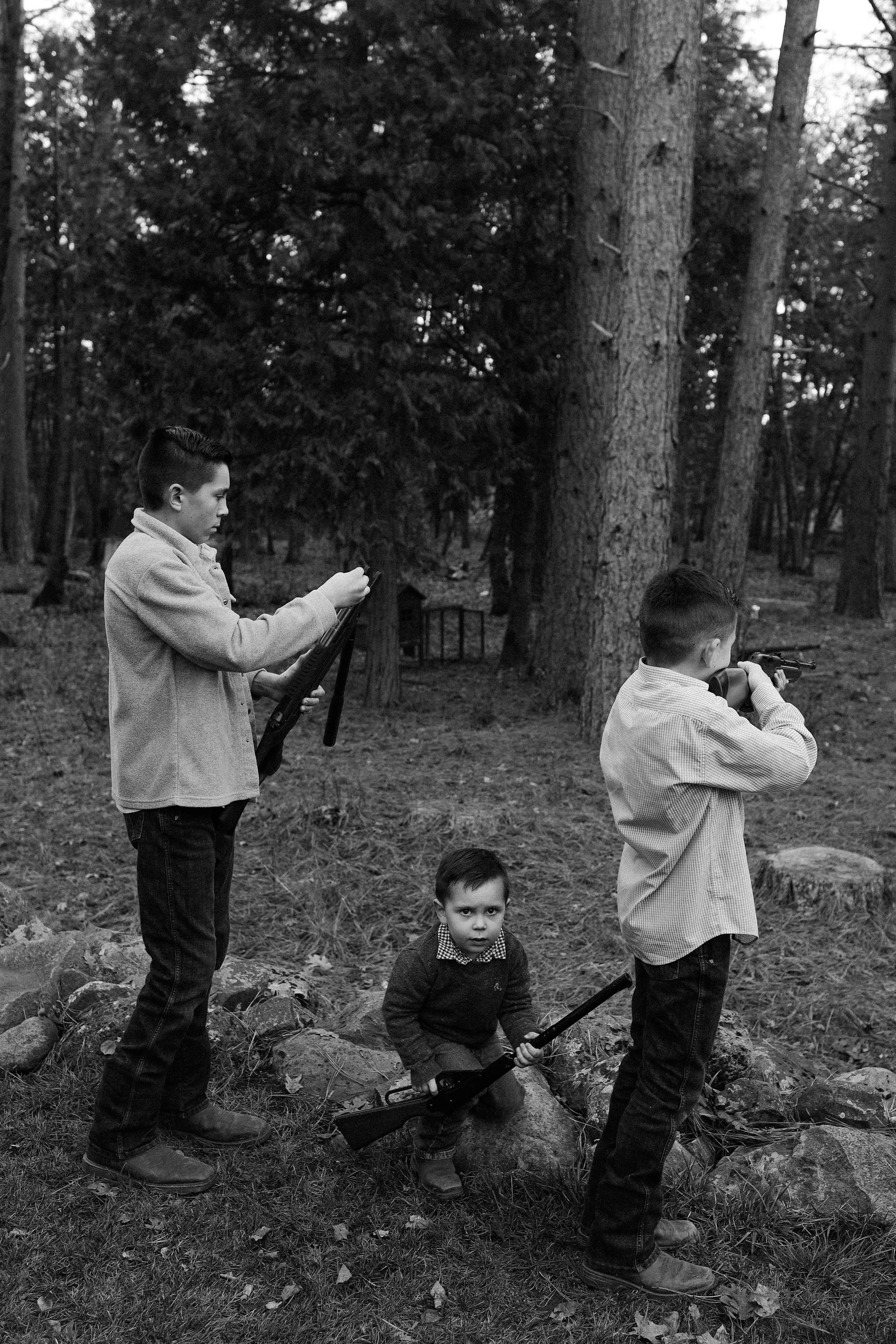 A black and white photo of four boys in a forested area, with two boys holding rifles, one standing and engrossed in a phone, and a small child sitting on the ground looking at the camera.