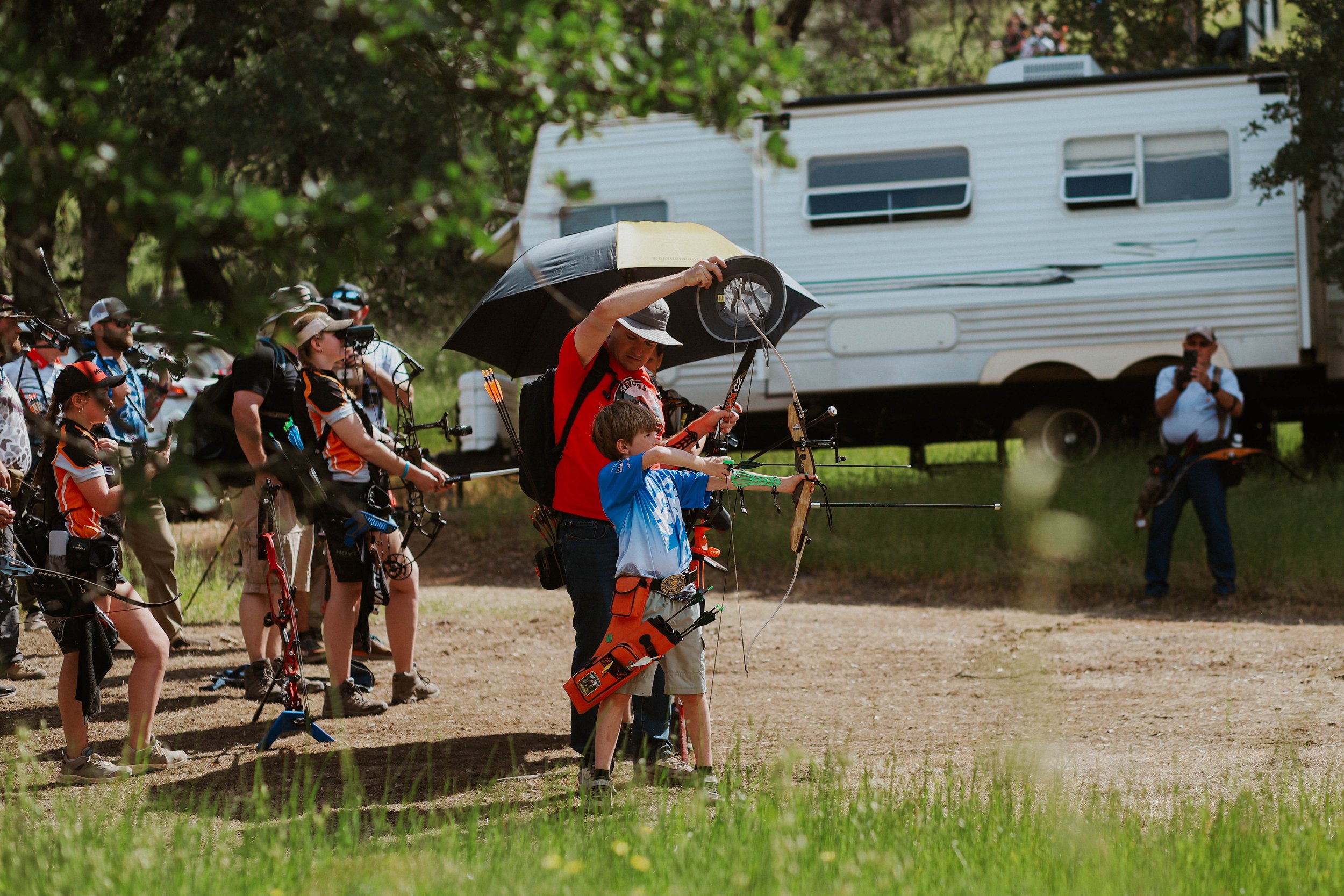 A group of people, including children and adults, participating in an outdoor archery activity. Some are aiming bows, and one adult male is holding a young boy's hand while adjusting his bow. There are trees and an RV trailer in the background.