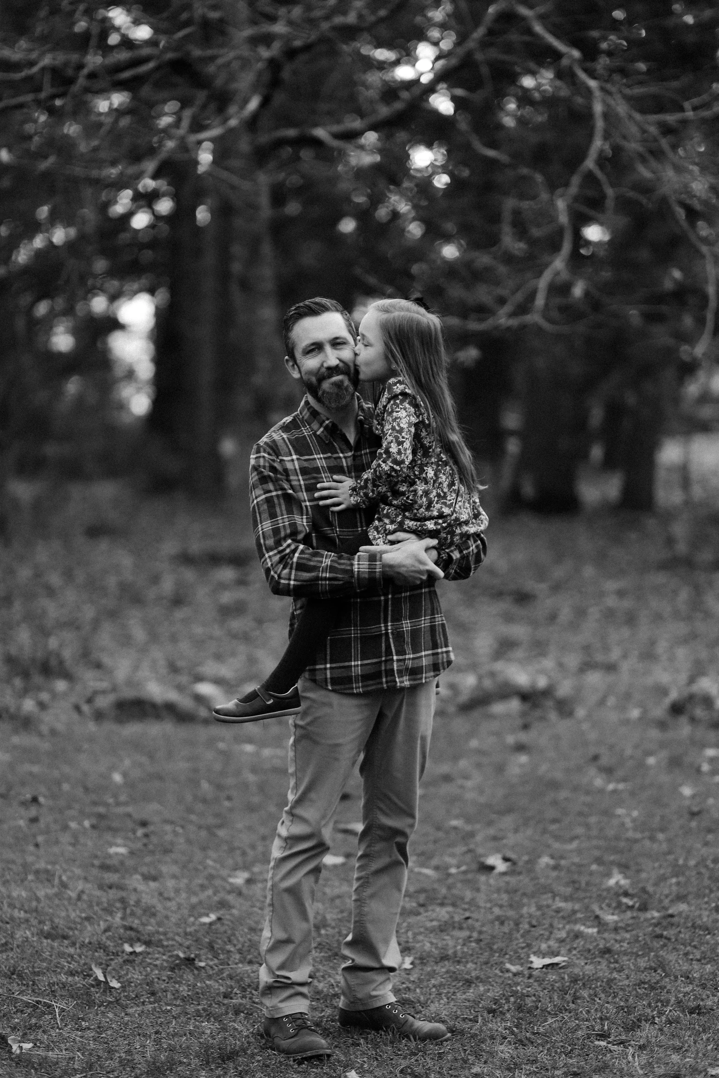A man holding a young girl in his arms outdoors, with trees in the background. The image is black and white.