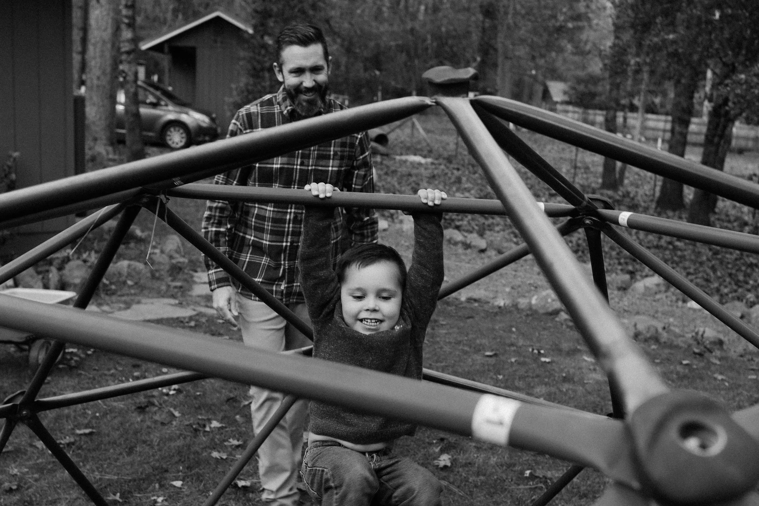 A man and a young child playing on a jungle gym outdoors, black and white photo, trees and house in background.