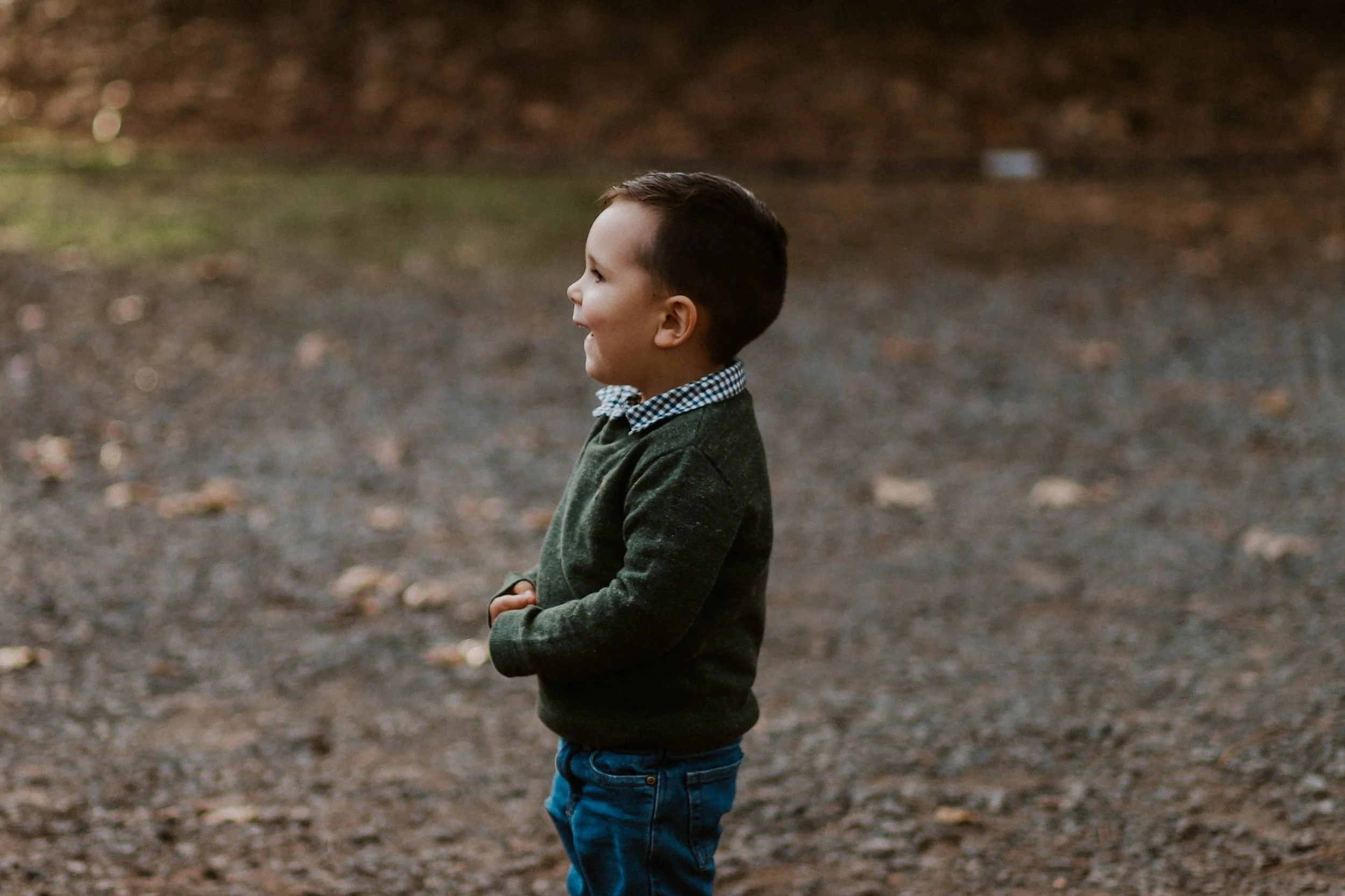 A young boy standing outdoors on a dirt ground, smiling and looking to his right side, with a blurry background of trees and fallen leaves.