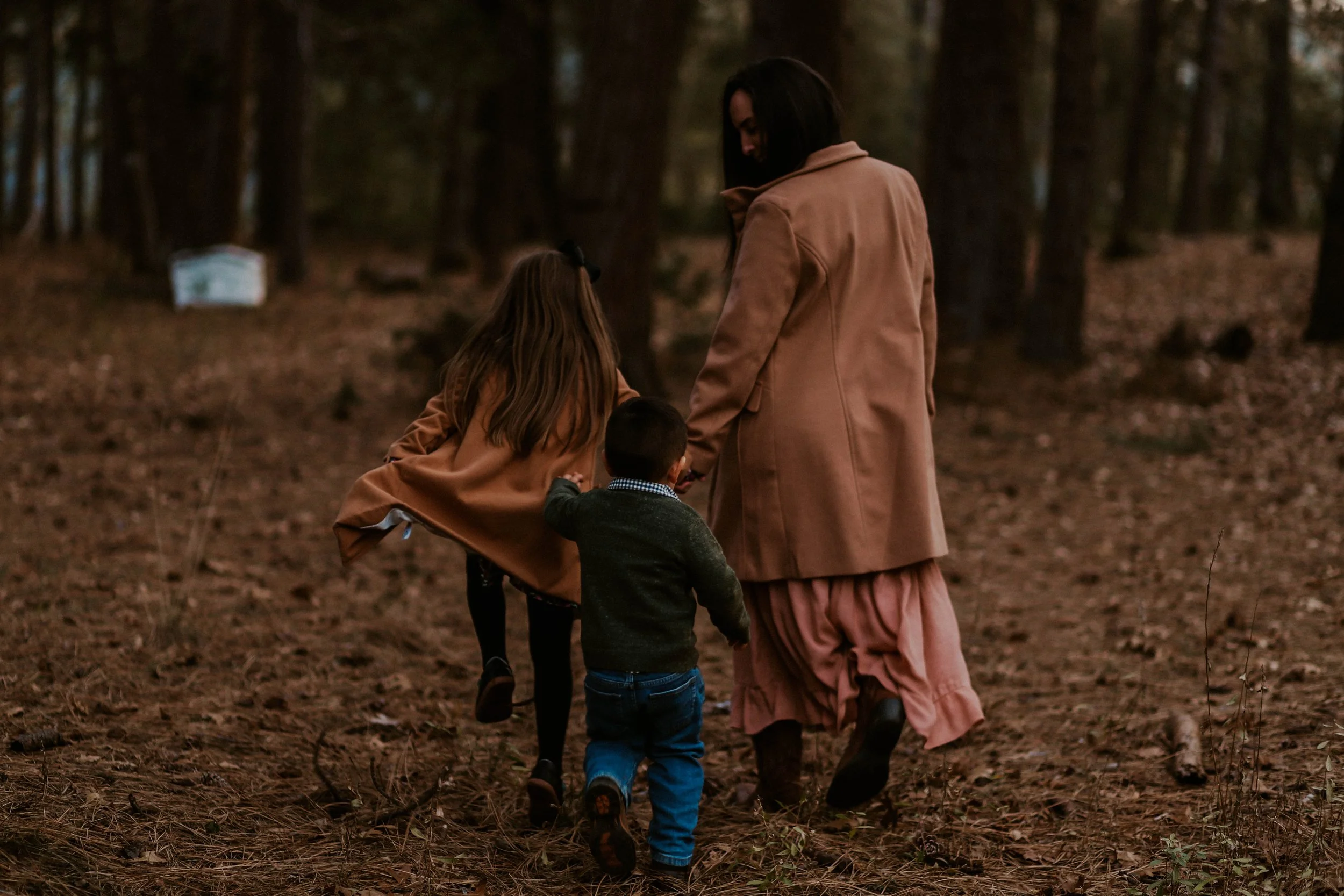 A woman and two children walk through a wooded park, holding hands, during the daytime. The woman is wearing a tan coat and a pink skirt, and the children are dressed in dark clothing.