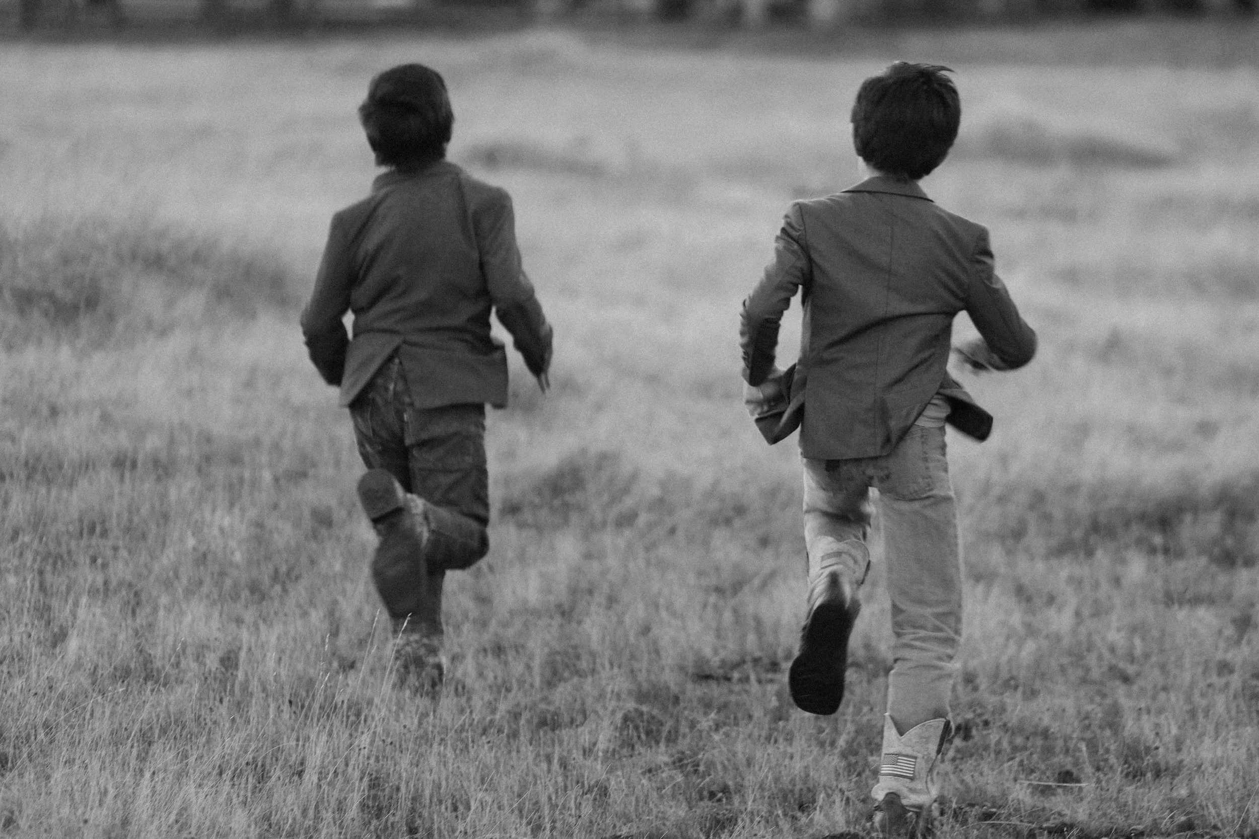 Two boys running through a grassy field with their backs to the camera, dressed in jackets and pants, on a cloudy day.