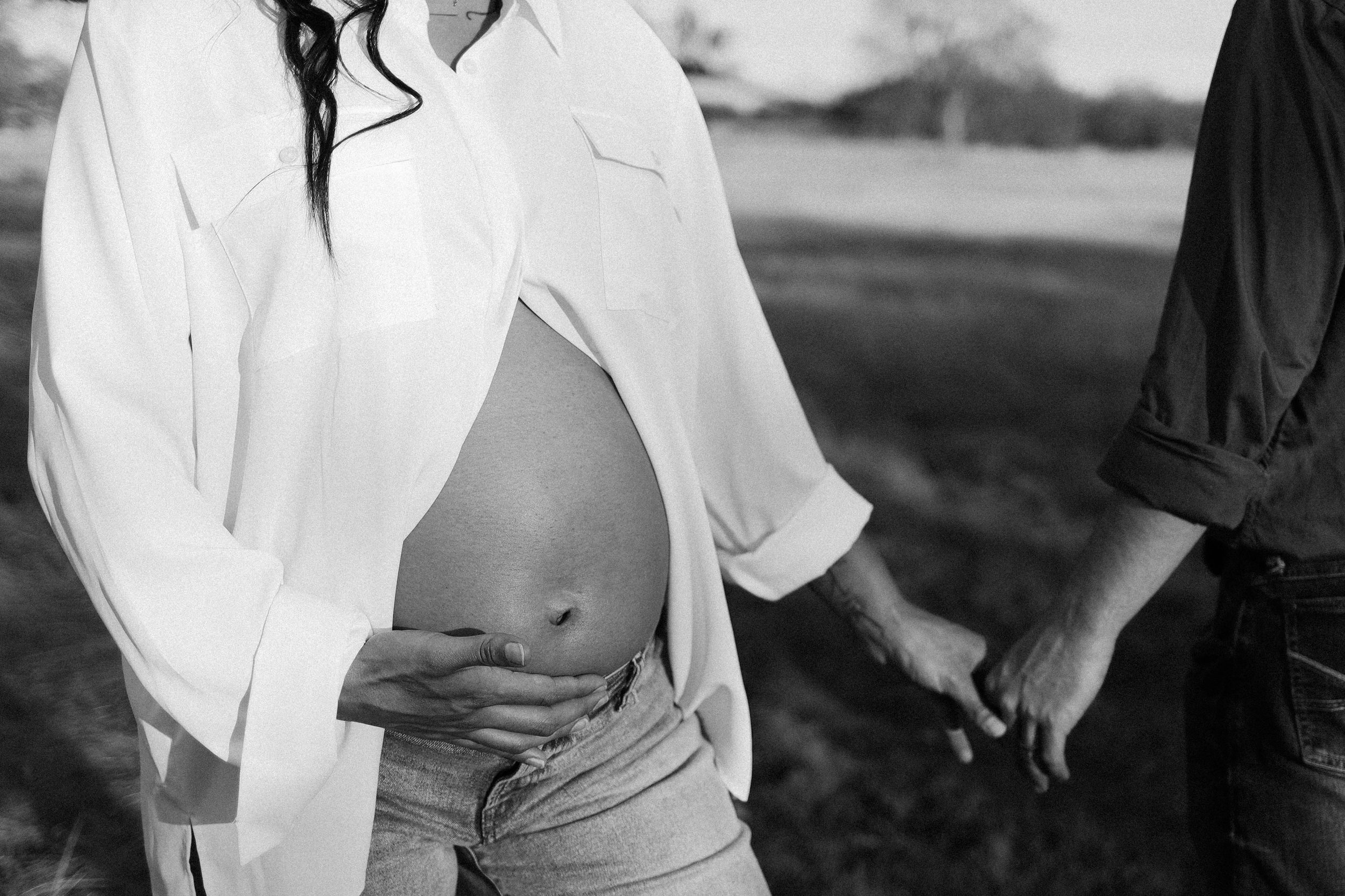 A woman with long, curly hair wearing a white shirt open at the front, revealing her pregnant belly, with hands resting on her hips, standing outdoors.