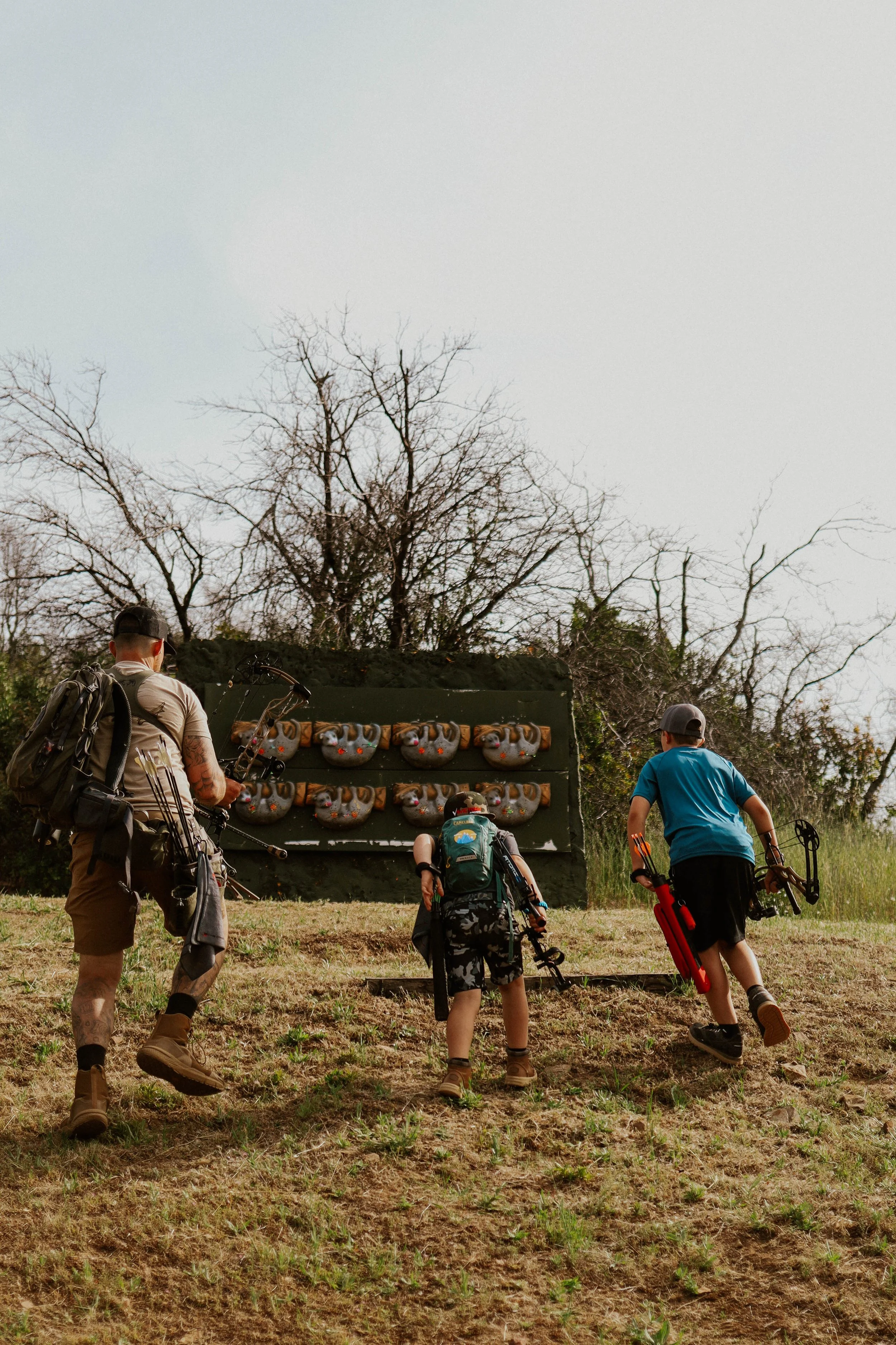 A group of four people, three children and an adult, heading towards a rock climbing wall with foam holds, in a grassy outdoor area with leafless trees in the background.
