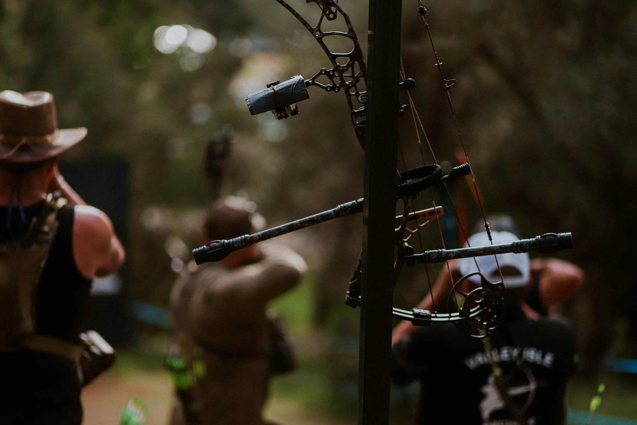 Archery target with a bow and arrow in focus, blurred background showing people in a wooded outdoor setting.