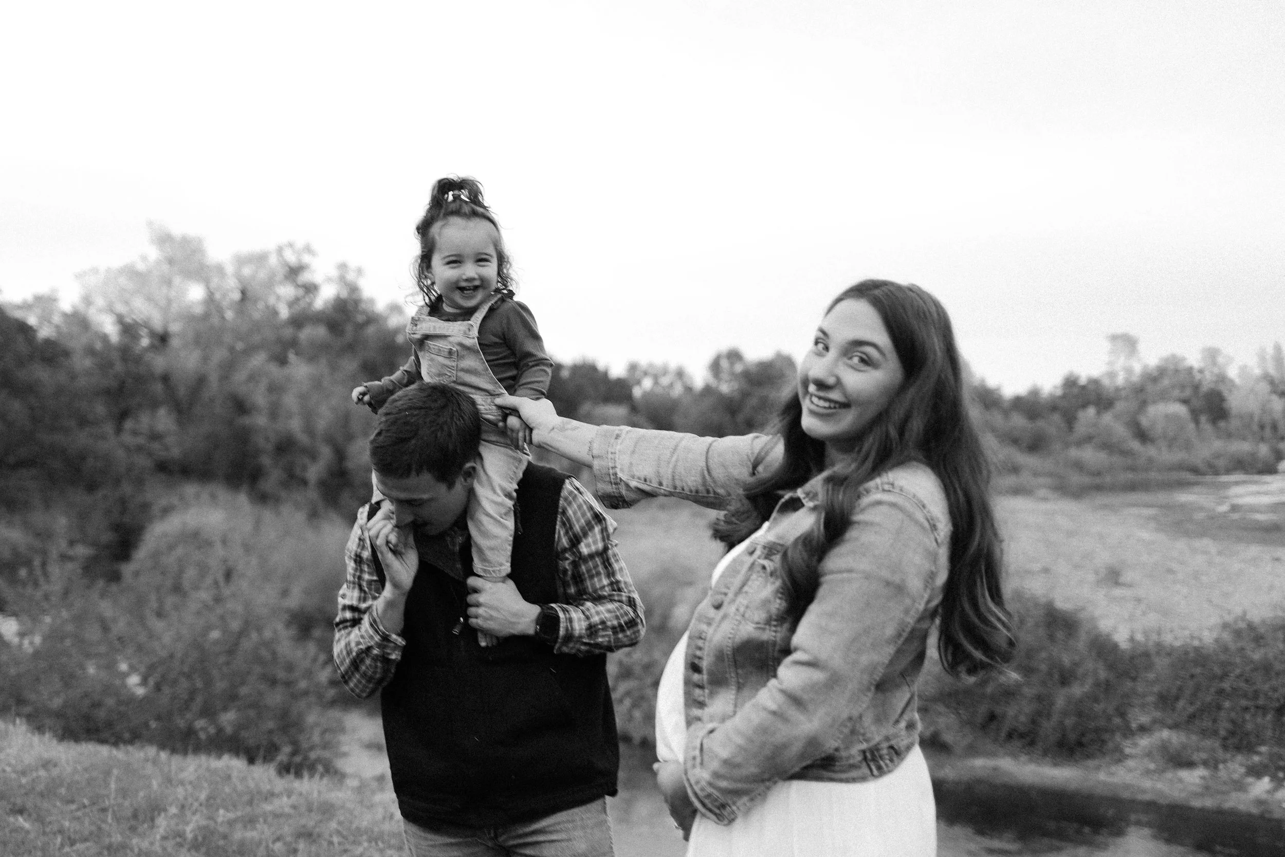 A father smiling and holding a young girl on his shoulders while the mother is in front of them outdoors. The girl is laughing, and all three appear happy in a natural setting.