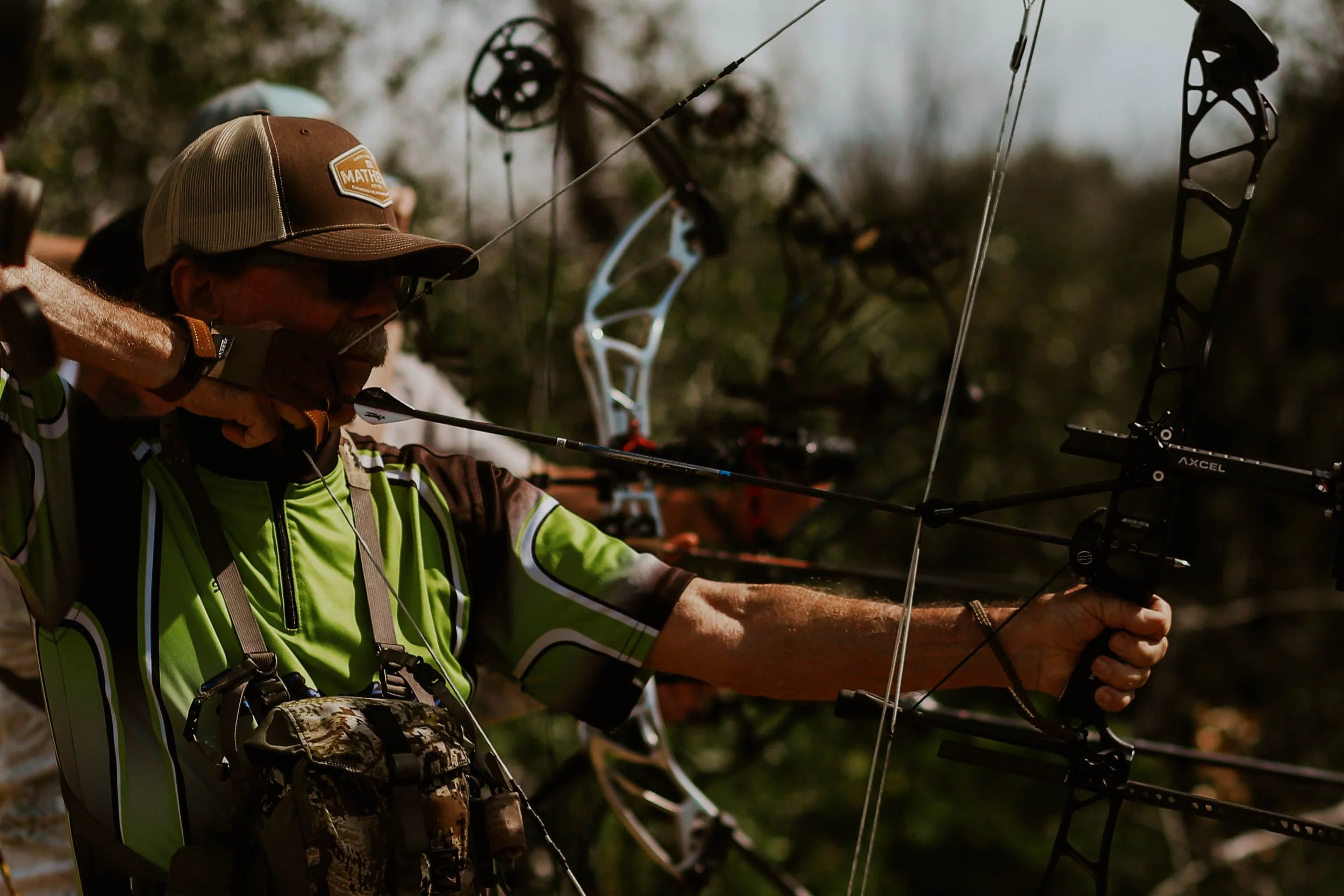 A person with a brown and beige cap and sunglasses is aiming a compound bow, preparing to shoot. The person is dressed in a green and black sports shirt, and there are multiple other bows in the background.