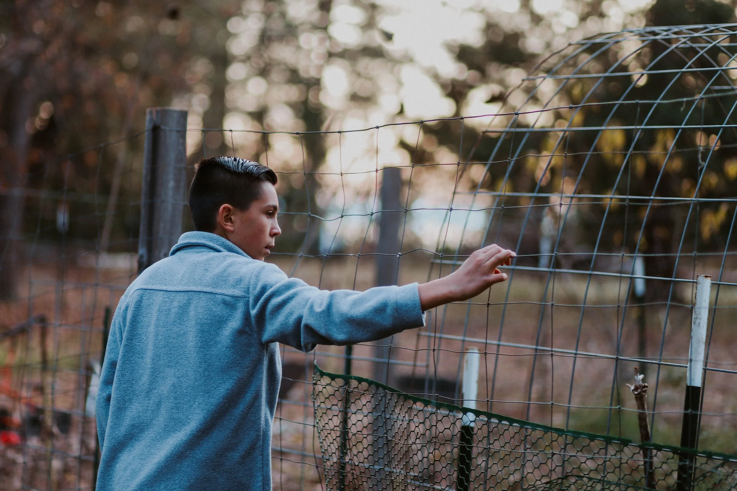A young person in a blue hoodie is looking at a wire fence, possibly inspecting or trying to repair it, in an outdoor setting with trees in the background during dusk.