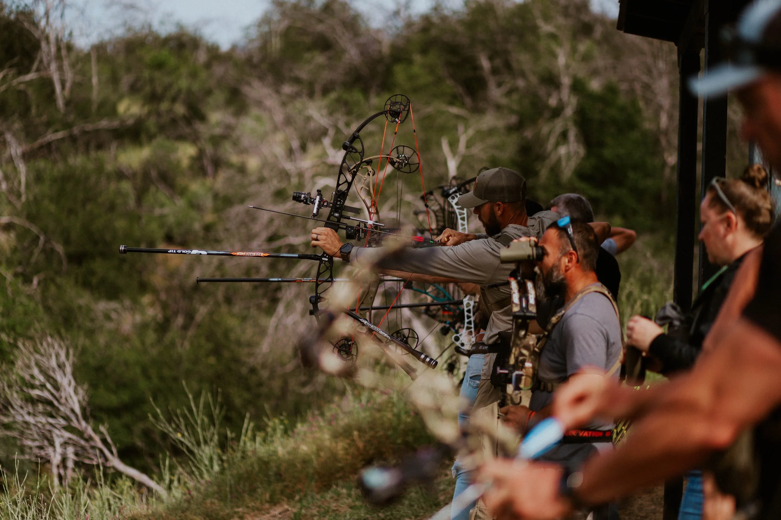 A group of archers practicing archery outdoors, aiming their bows at targets in a natural setting with trees in the background.