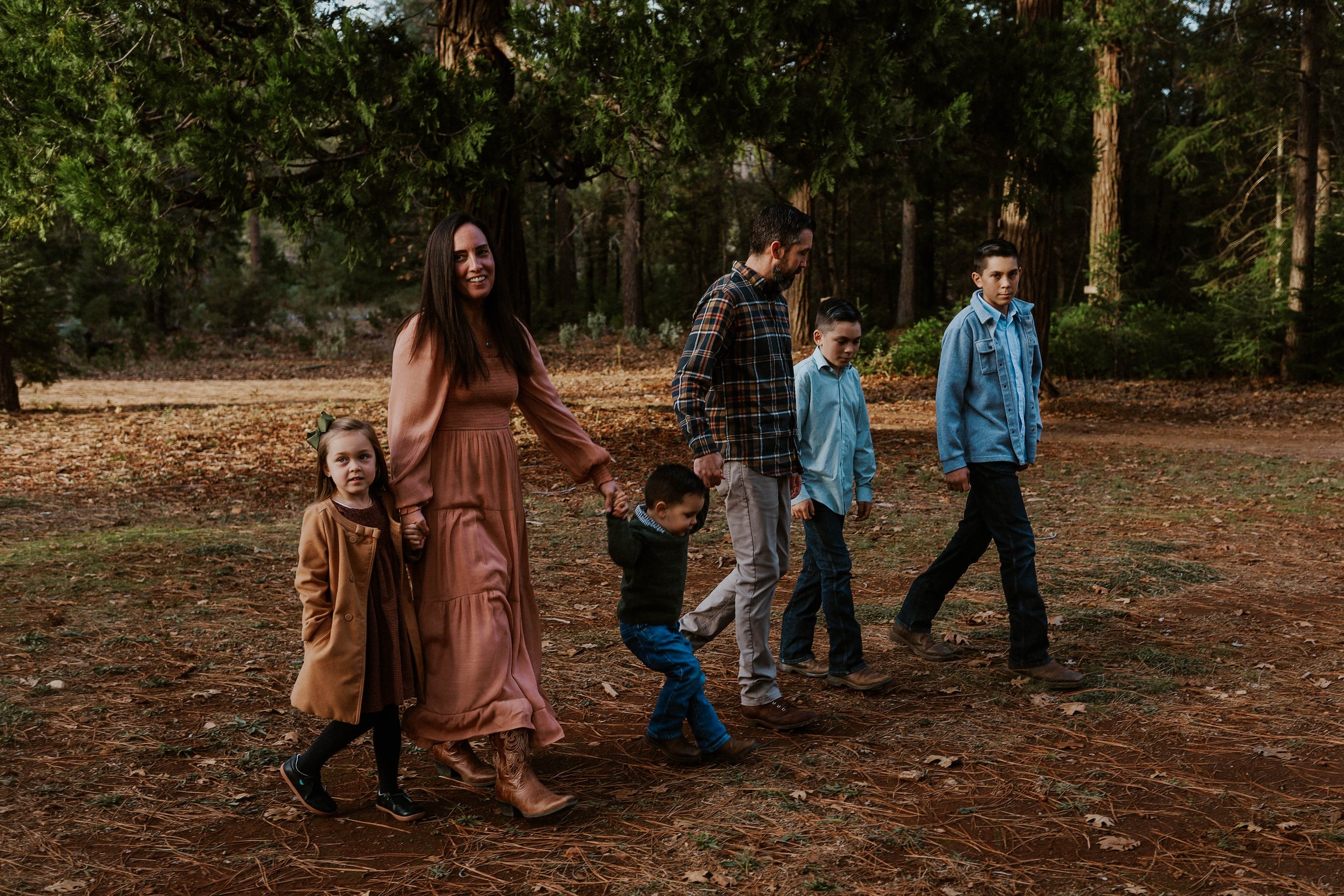 A group of people walking through a wooded area, consisting of a woman, three boys, and a girl, all dressed casually and holding hands.