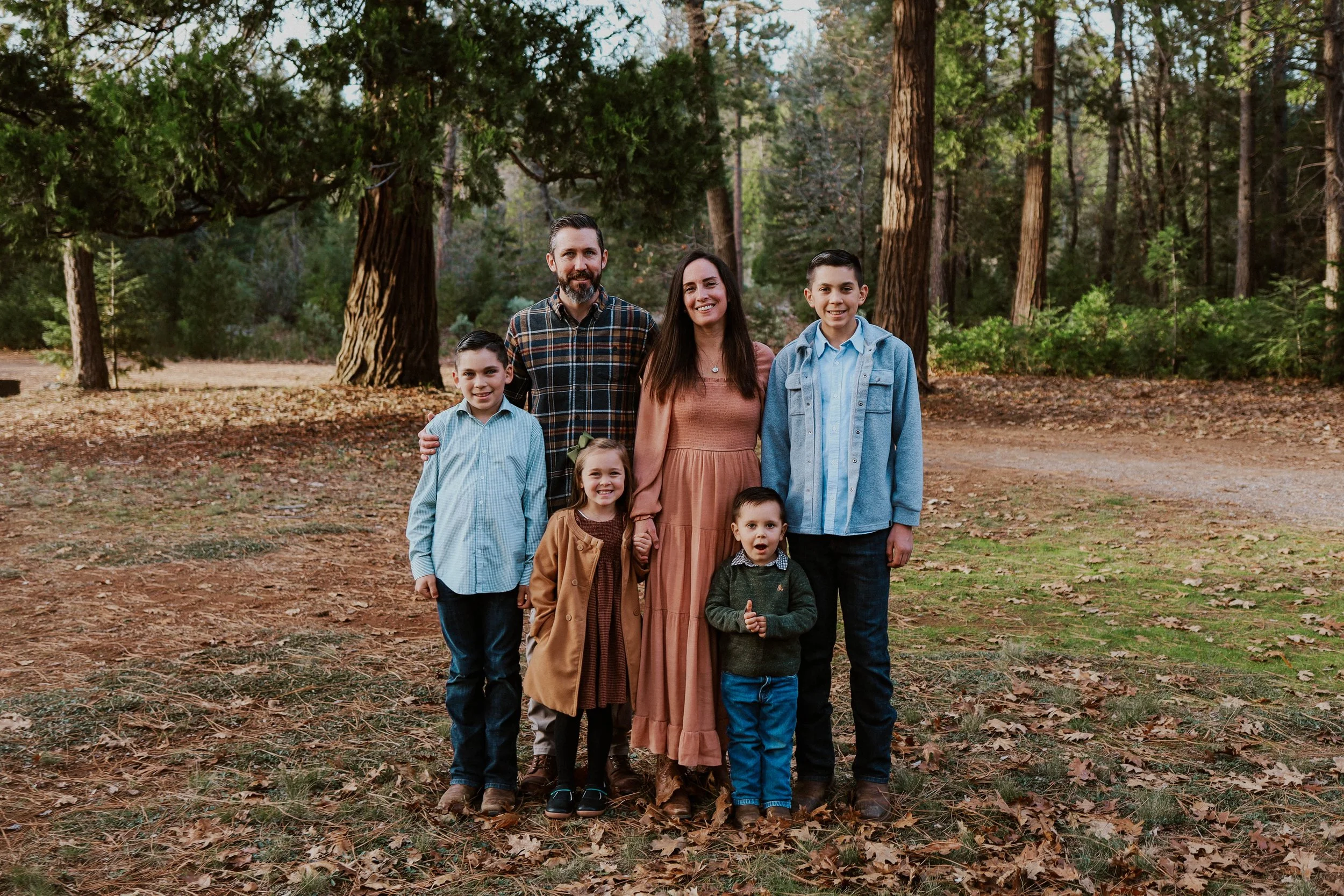 A family of eight people standing outdoors in a wooded area with tall trees and fallen leaves, smiling at the camera.