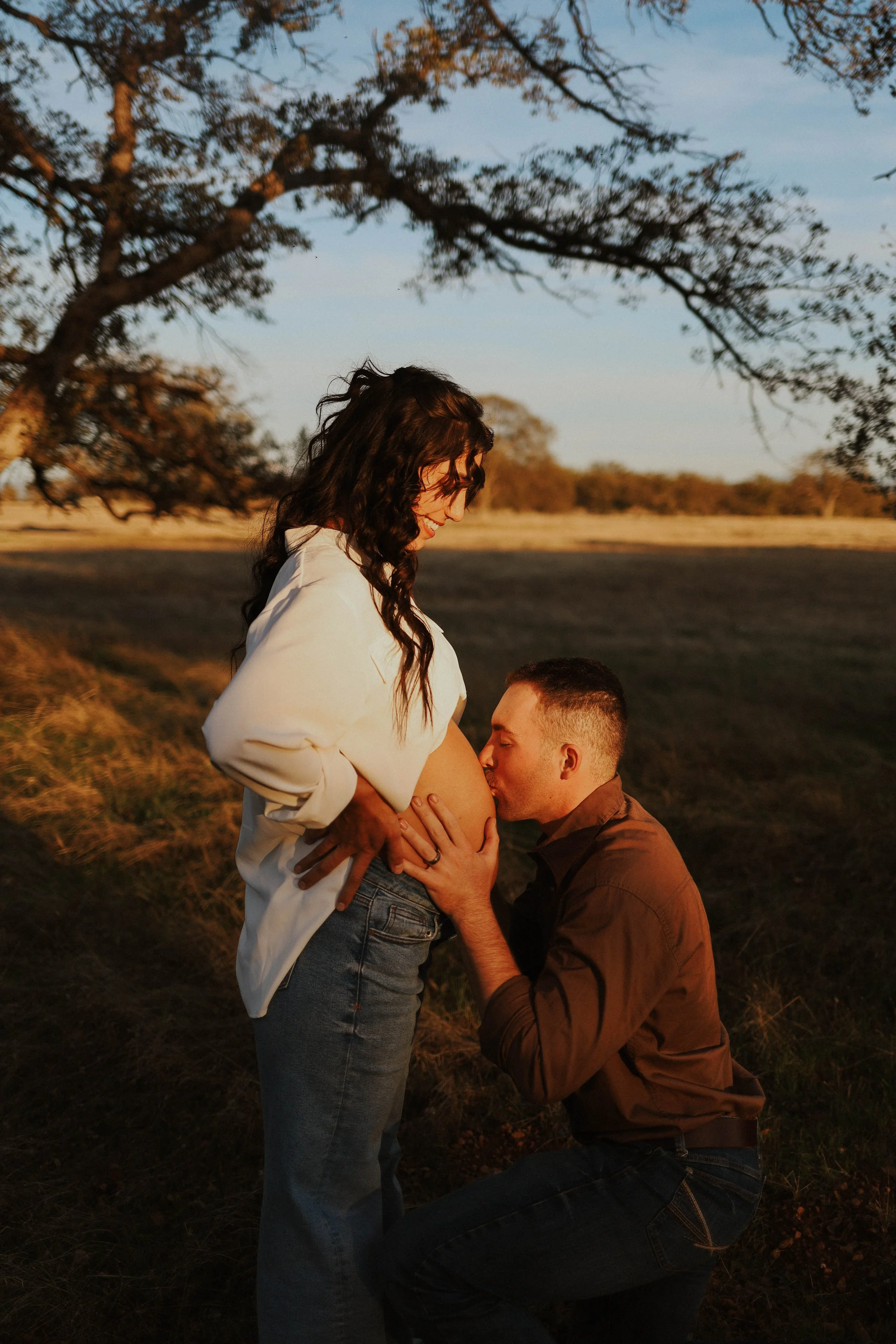 A couple during a pregnancy photo shoot outdoors at sunset. The woman is smiling and looking down at her pregnant belly while the man kneels in front of her, holding her belly and kissing it. The background features trees and a field.