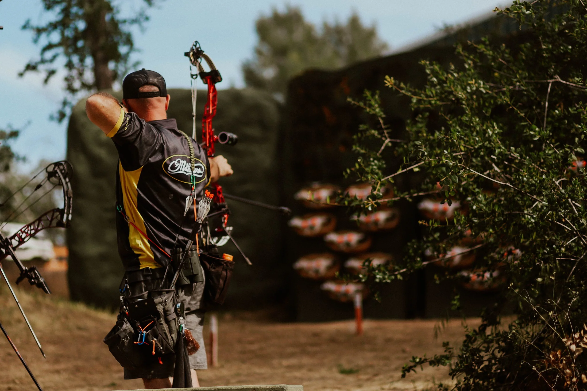 A person using a compound bow in an outdoor setting, aiming at a target, with trees and a shaded area in the background.