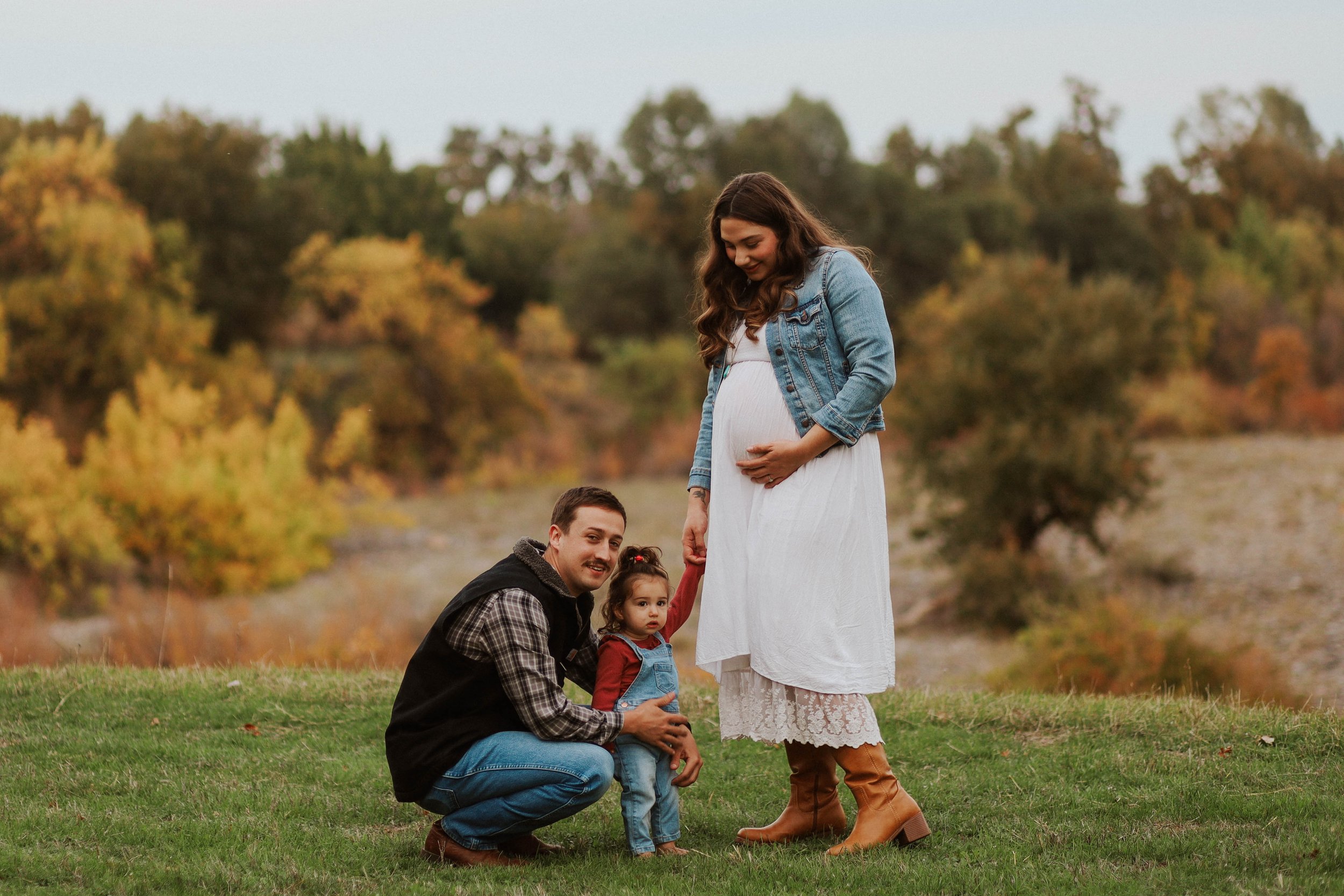 Family outdoor photo with woman in white dress and denim jacket, man in black jacket, and a child, standing on grass with autumn trees in background.