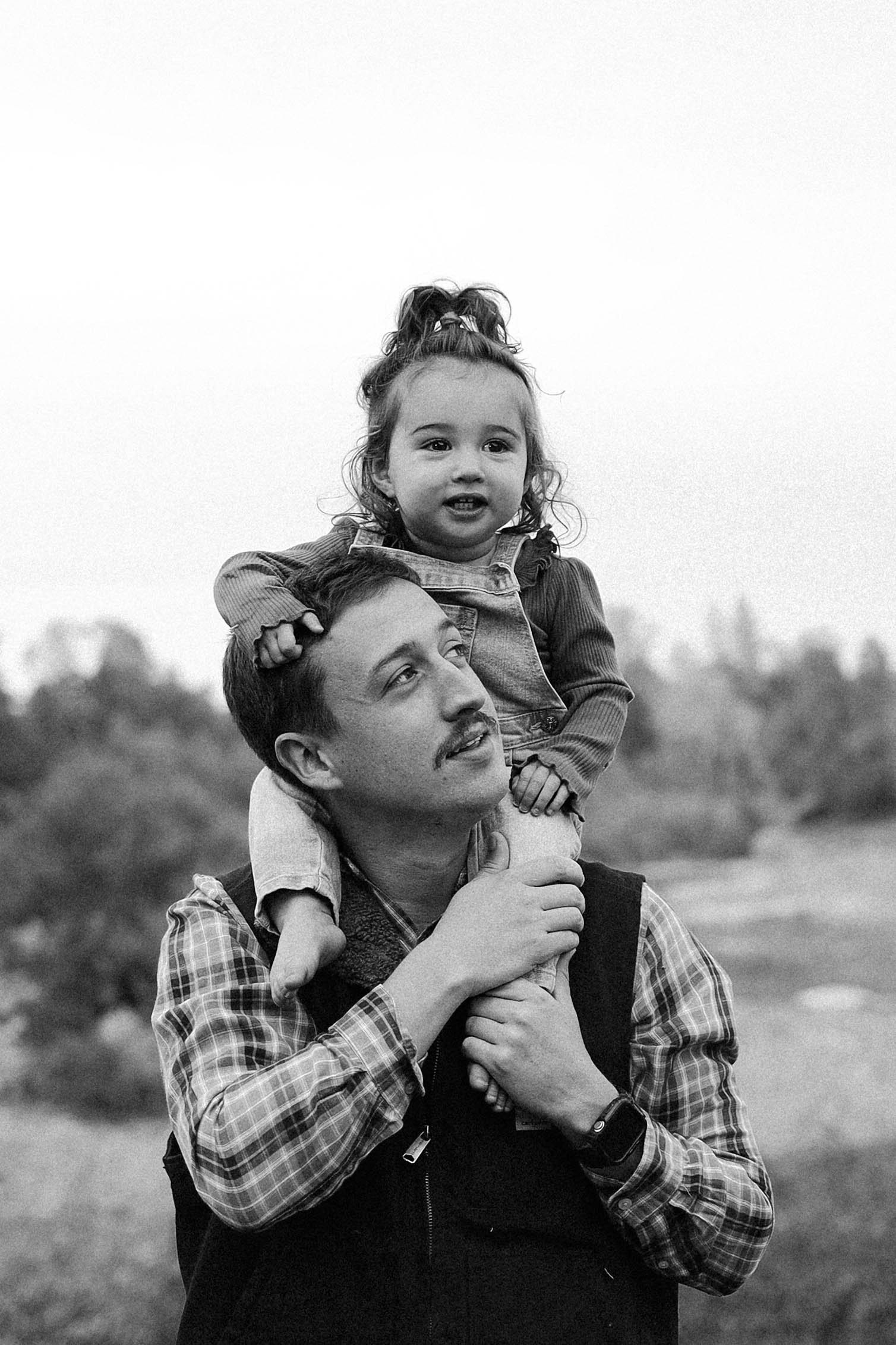 A man is holding a young girl on his shoulders outdoors, with both smiling and looking happy. The background is blurred with trees, indicating a natural setting.