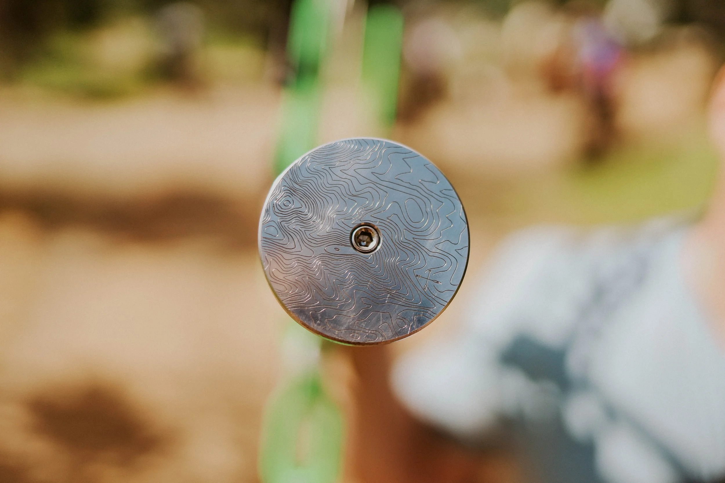 Close-up photo of a round metallic object with a detailed topographical map pattern, mounted on a green support, with a blurred background of grass and plants.