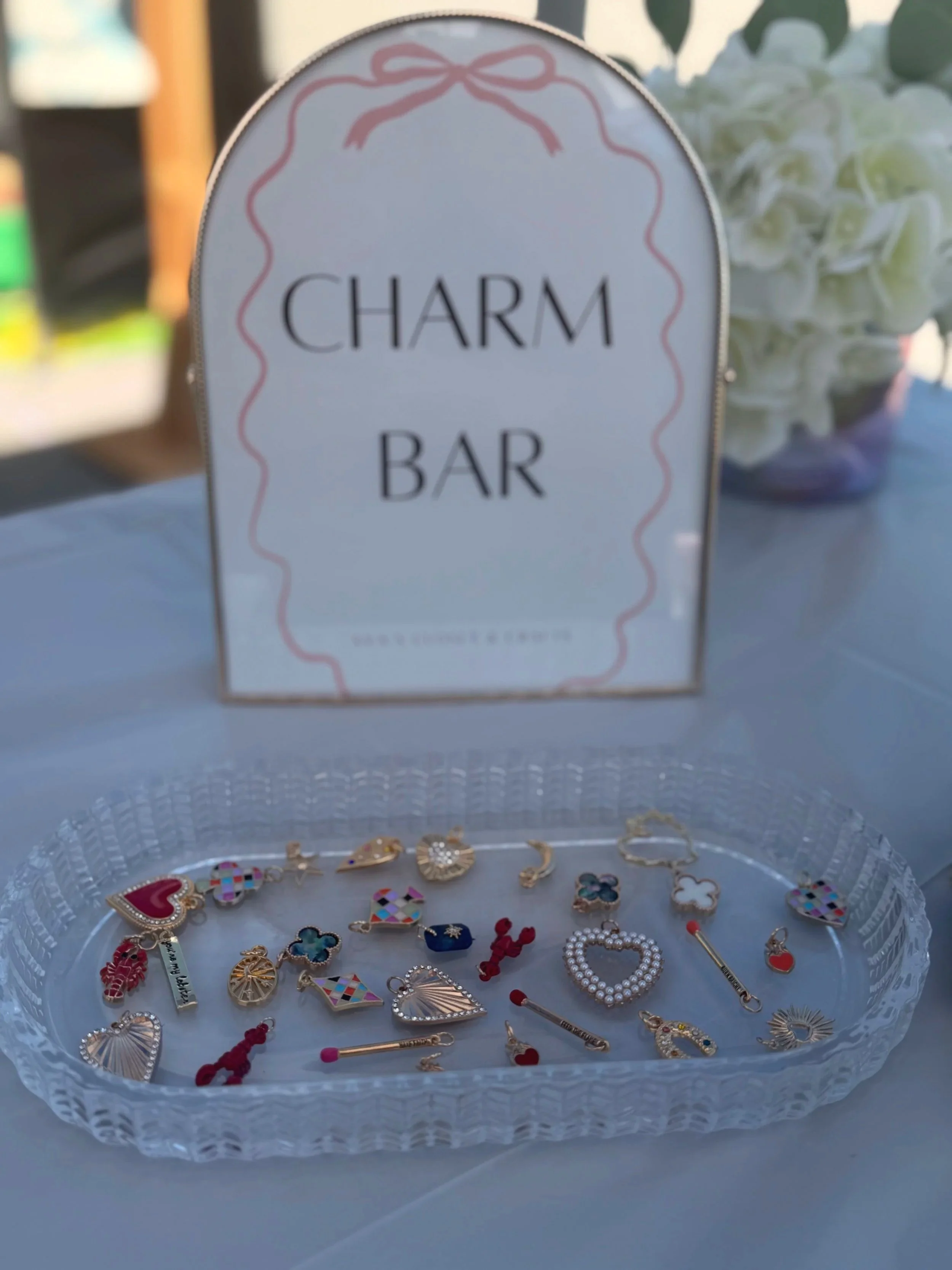 A display of various colorful earrings and jewelry pieces in a clear plastic tray, with a sign that reads 'Charming Charm Bar' in the background.