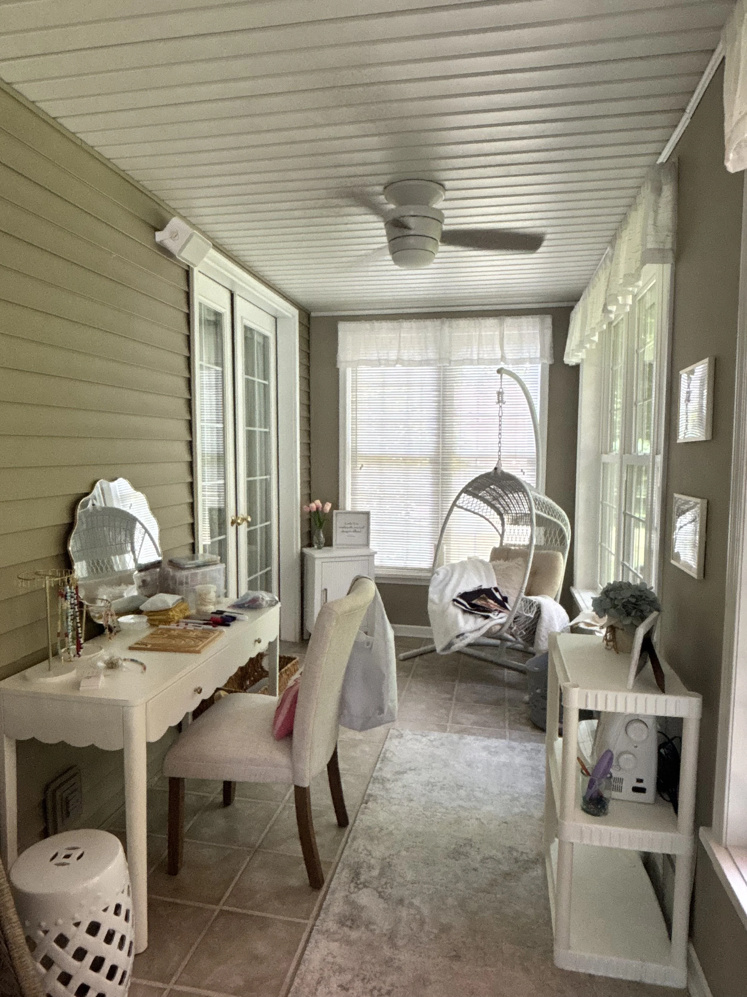 A cozy sunroom with beige vinyl siding, large windows with white blinds, and white furniture including a chair and side tables. Decor includes a hanging wicker chair, a small table with a plant, and various accessories. The room is softly lit with natural light.
