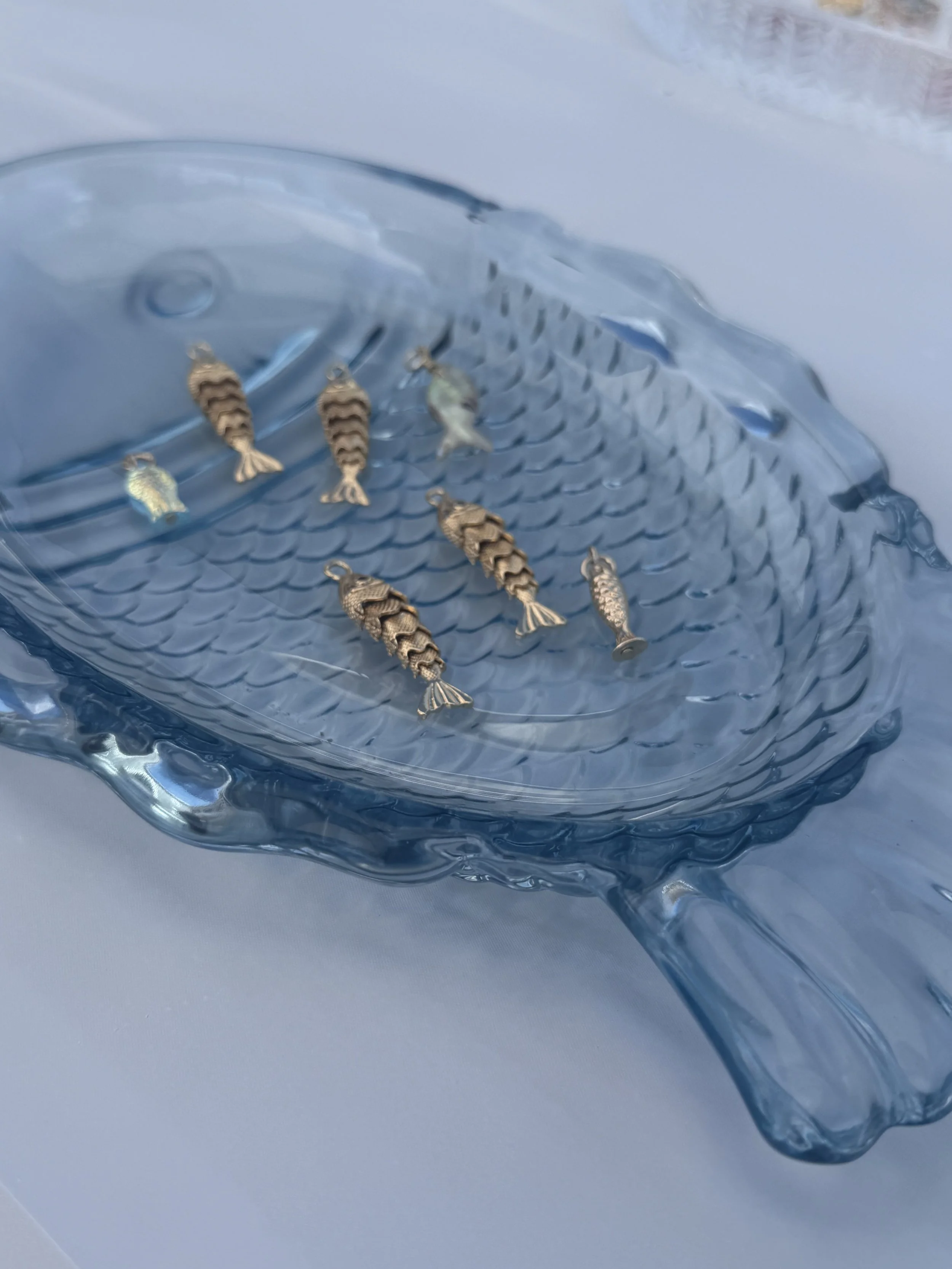 A clear glass fish-shaped container filled with small, gold-colored fish-shaped charms or pendants with textured bodies and fins.