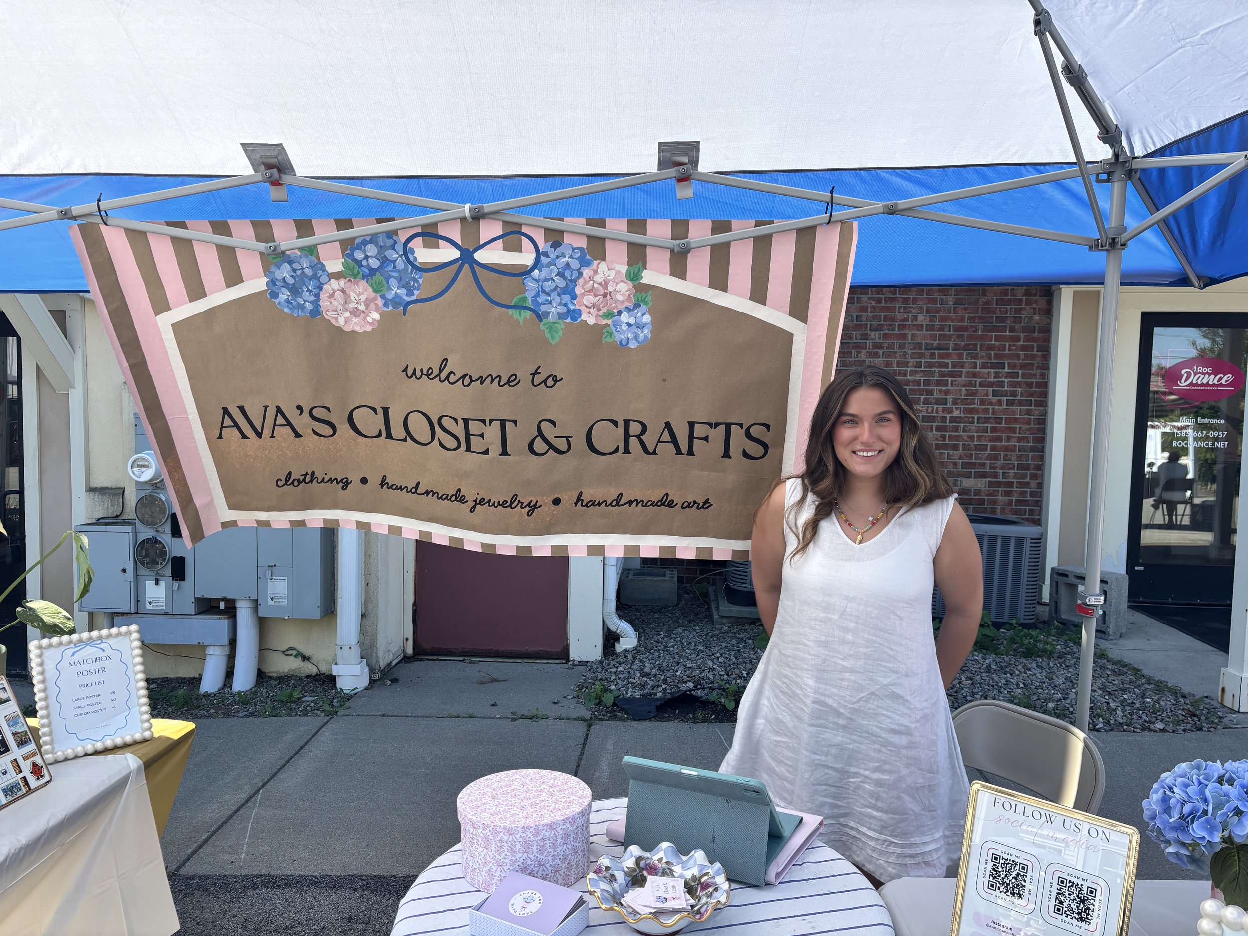 A young woman standing outside in front of a booth for Ava's Closet & Crafts with a sign that reads "welcome to Ava's Closet & Crafts, clothing, handmade jewelry, handmade art." The booth is under a blue canopy, with a table displaying various items, and a woman smiling at the camera.
