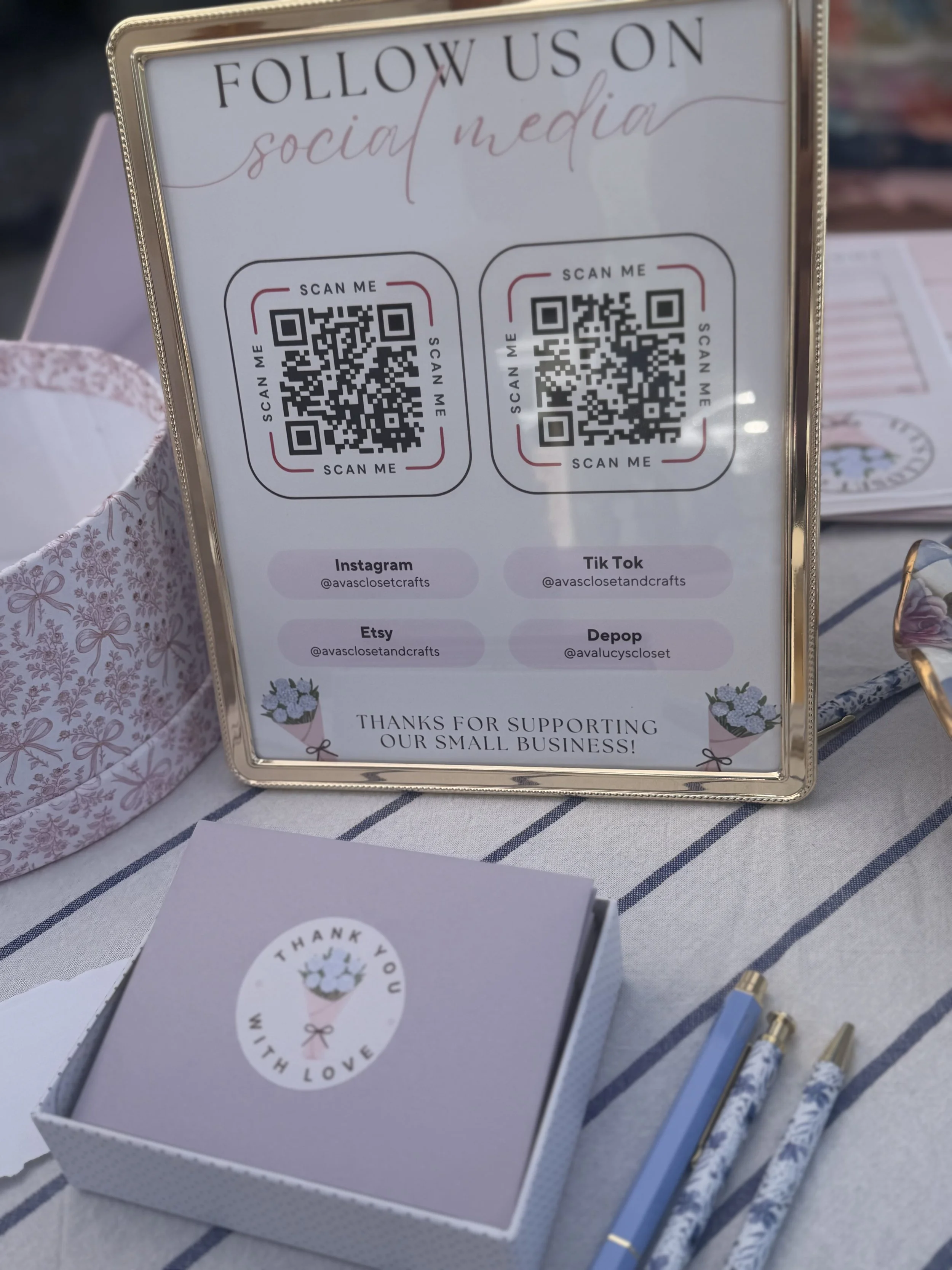 Sign with social media QR codes for a small business, including Instagram, TikTok, Etsy, and Depop, placed on a table with decorative items and pens.