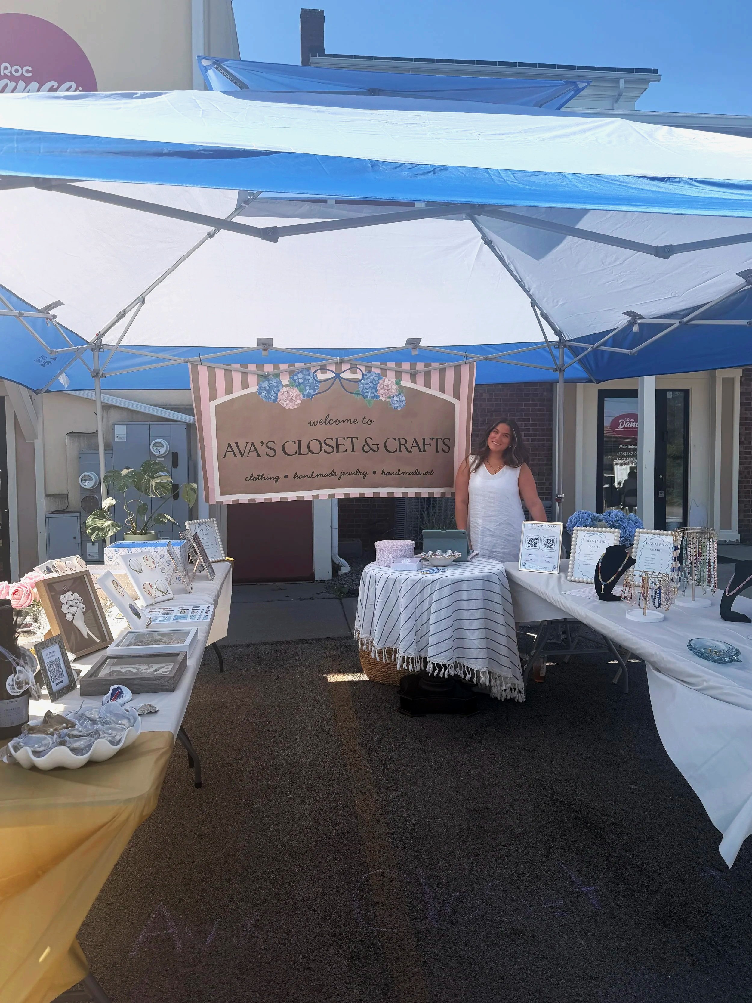 Outside craft booth with jewelry and accessories on display, awnings overhead, a woman smiling behind the table, and a large sign for "Ava's Closet & Crafts."