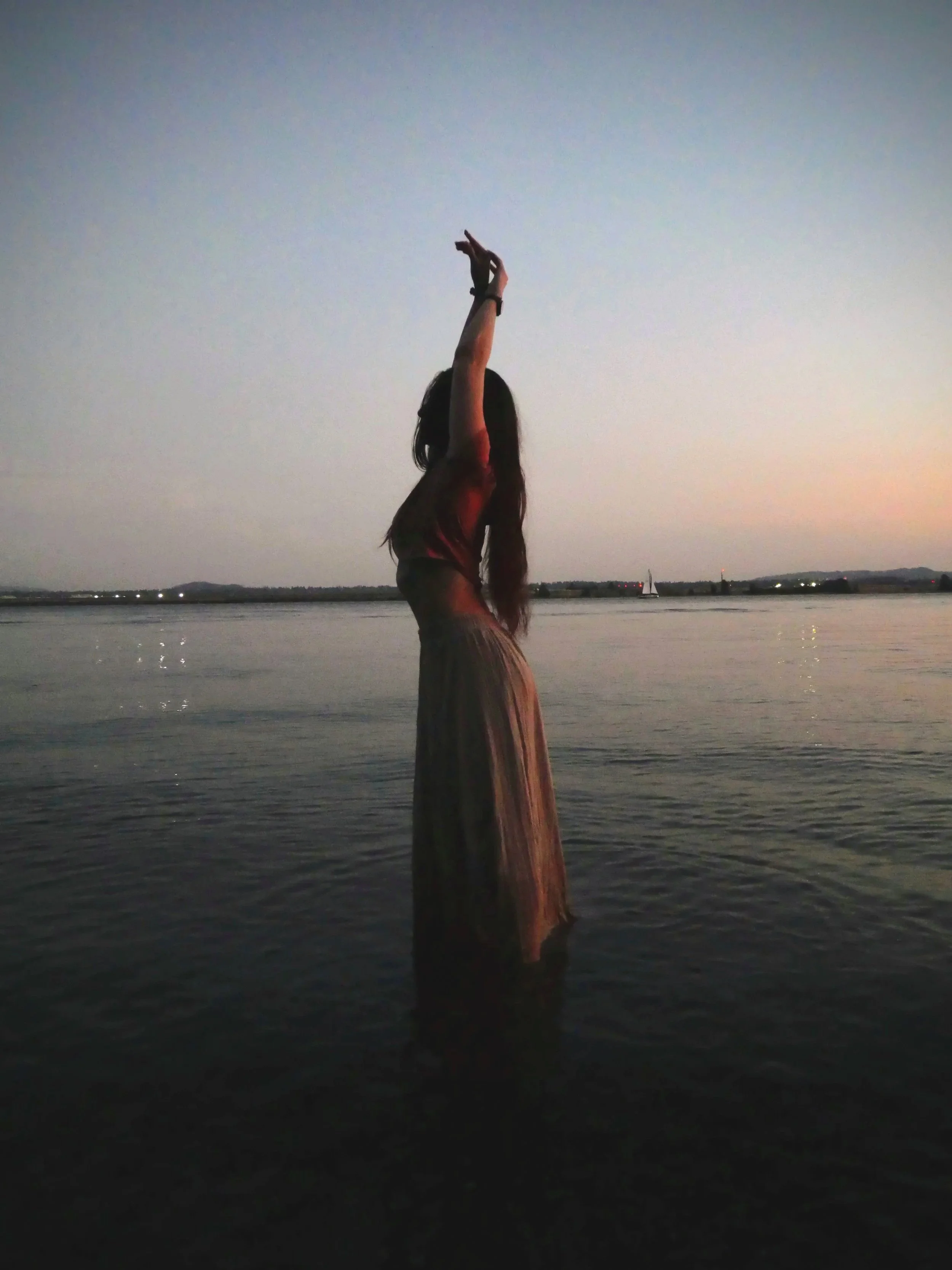 A woman with long hair standing on a body of water with her arms raised above her head, looking toward the sky during sunset or dusk, with a sailboat visible in the distance.