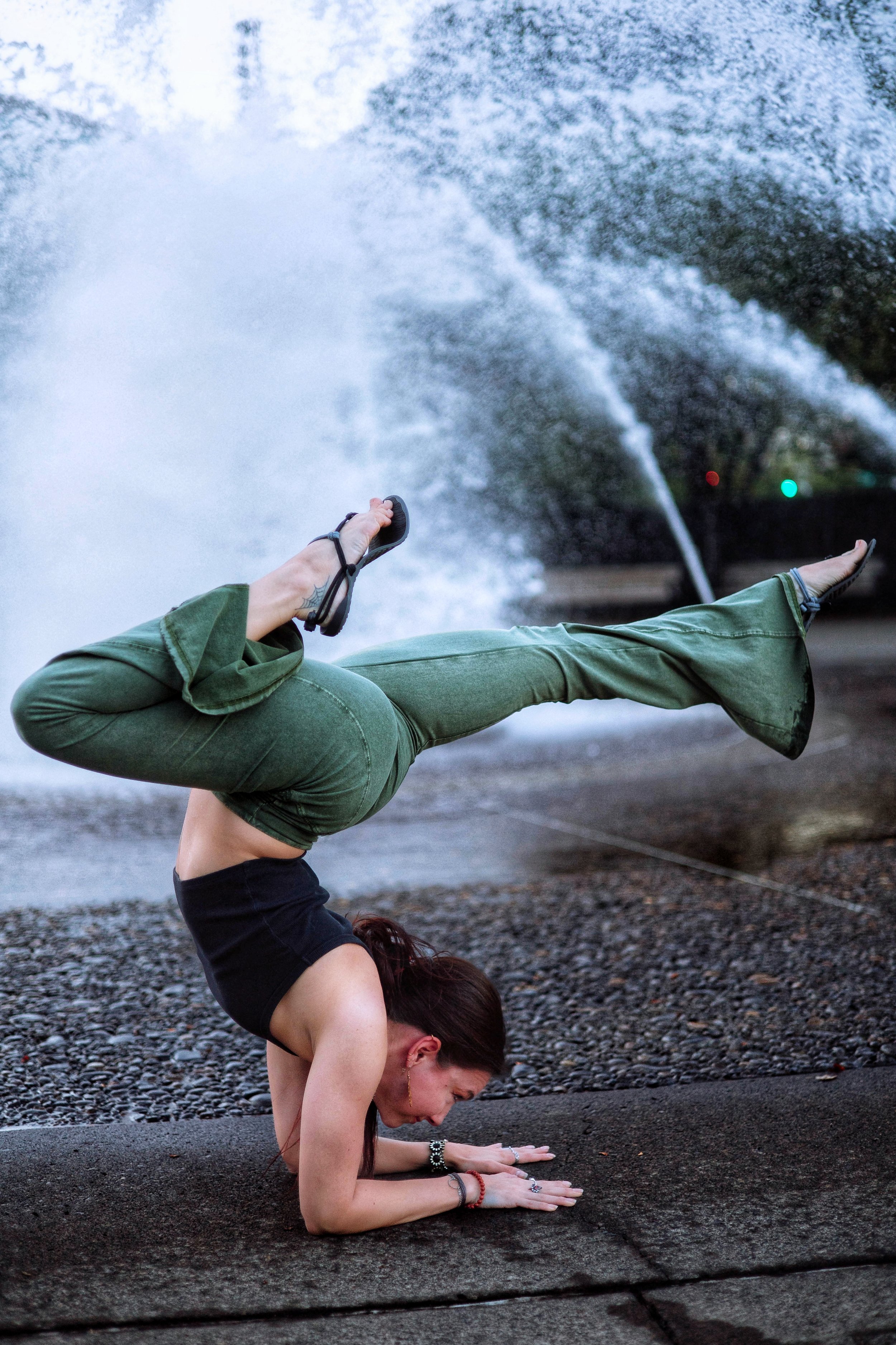A woman performing a hand-stand yoga pose on a gravel surface outdoors, with stream water shooting from a fountain behind her. She is wearing a black sleeveless top, green pants, and has long hair and jewelry on her wrists.