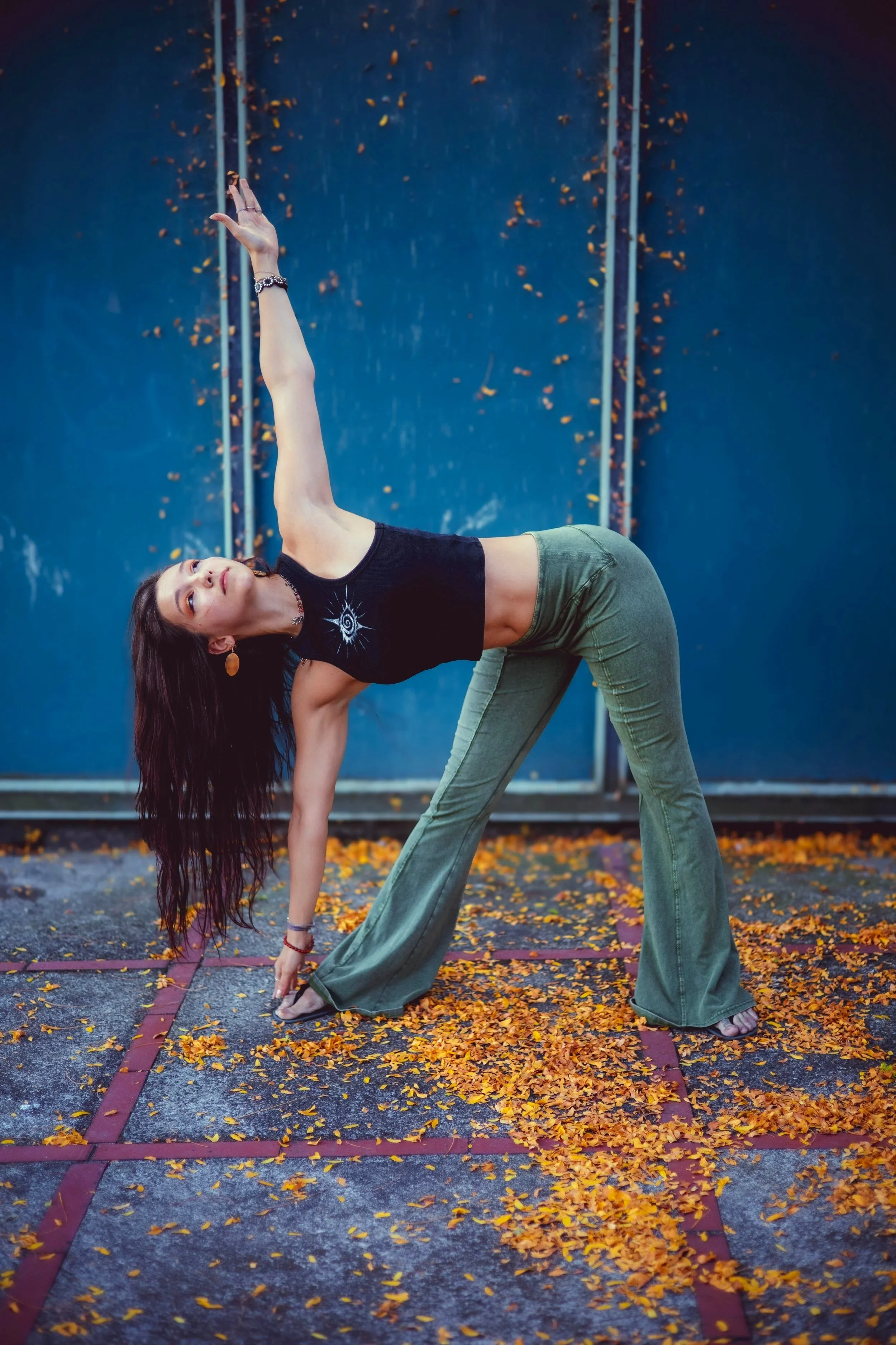 A young woman with long dark hair doing yoga outdoors on a marked section of pavement covered with fallen yellow autumn leaves. She is in a bent-over pose with one arm reaching upward and the other touching the ground, wearing a black sleeveless top, green pants, jewelry, and earrings, against a blue background.