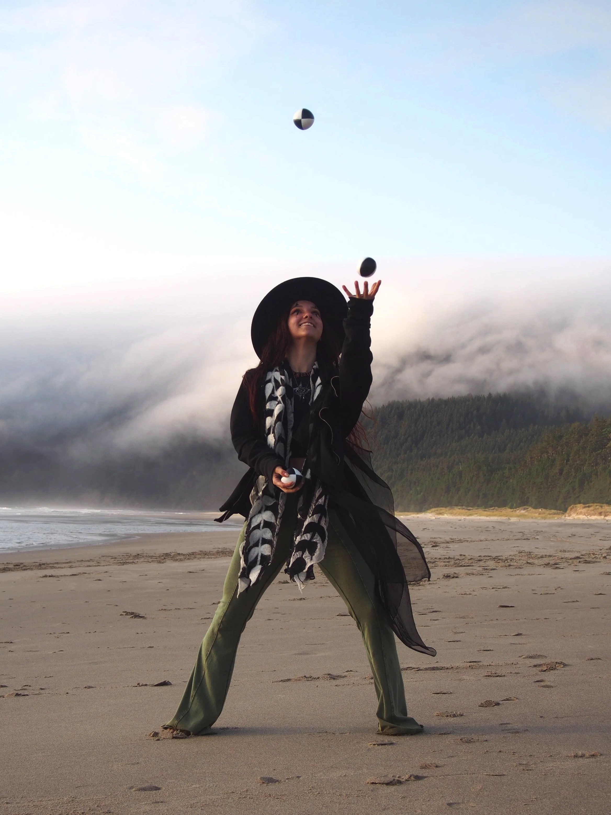 A woman on a beach with long pants, a black coat, a patterned scarf, and a wide-brimmed hat, throwing two toy black and white balls into the air with a forested hillside and cloudy sky in the background.