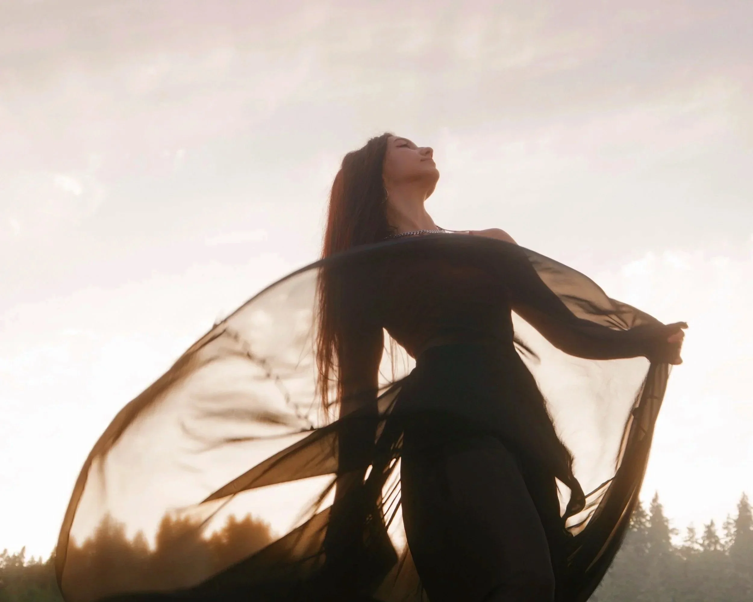 A woman with long dark hair wearing a black dress or top, standing outdoors with arms outstretched, looking up with eyes closed, in front of a cloudy sky during sunset or sunrise.