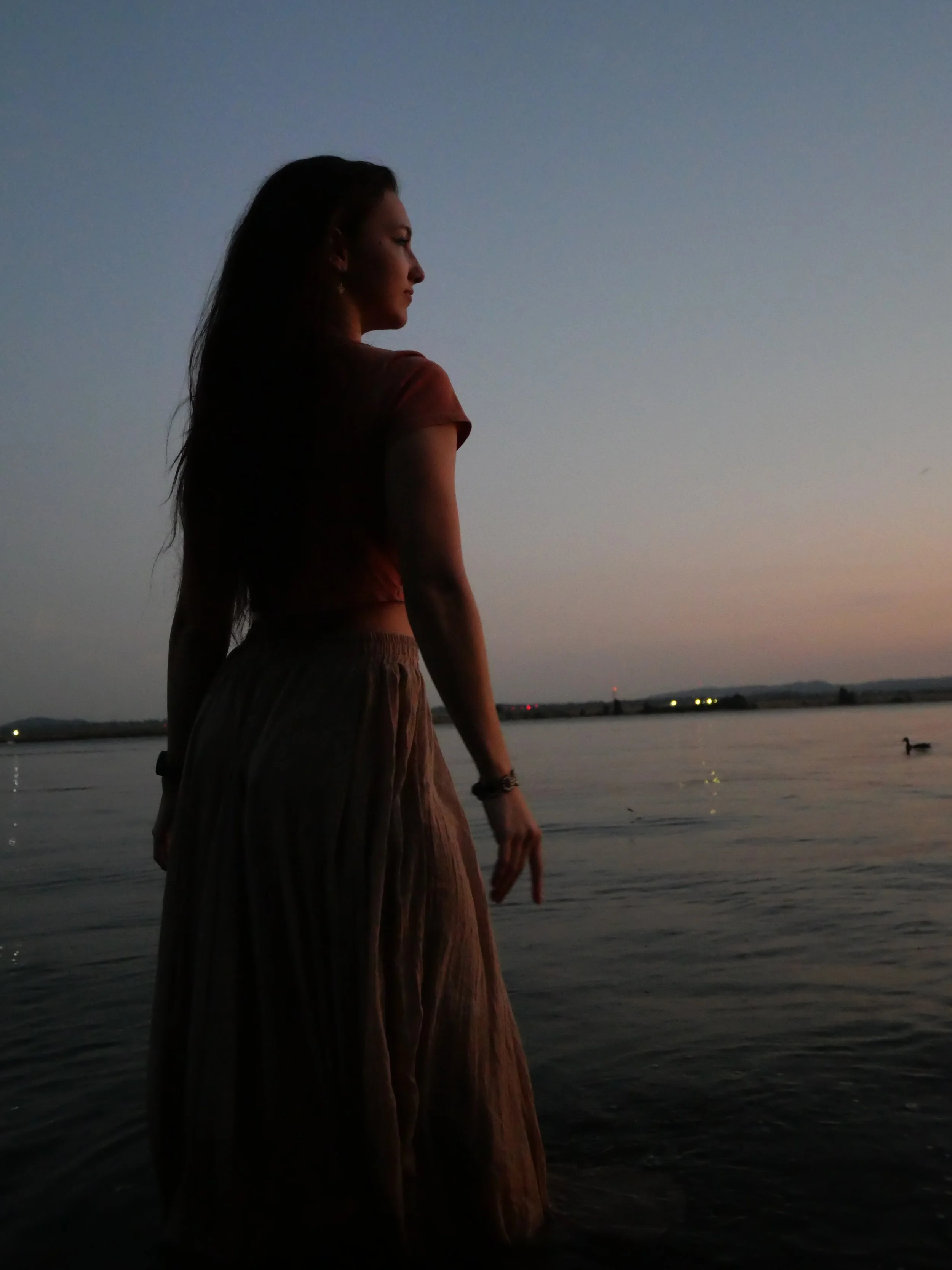 A woman standing by a body of water at dusk, with a small boat visible on the water and a distant shoreline with lights.