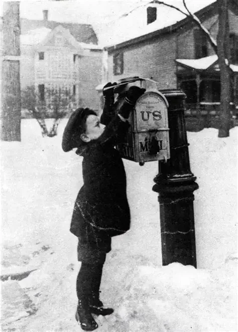 A young boy in winter clothing standing outside, mailing a letter into a USPS mailbox amid snow, with houses in the background.