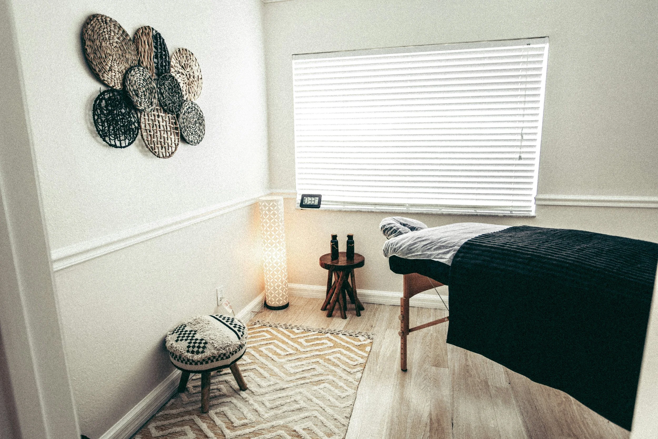 A cozy bedroom with wall decor, a window with blinds, a bed partially visible, a small side table with bottles, a lamp, a decorative ottoman, and a patterned rug.