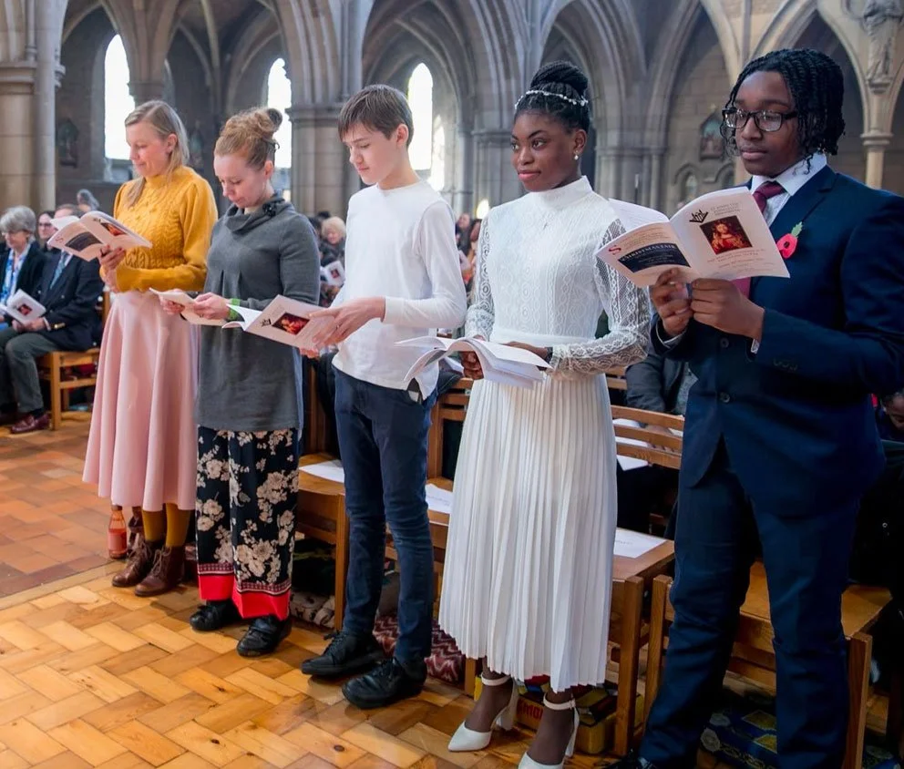 Children standing in a church, holding programs and participating in a ceremony. They are dressed in formal attire.