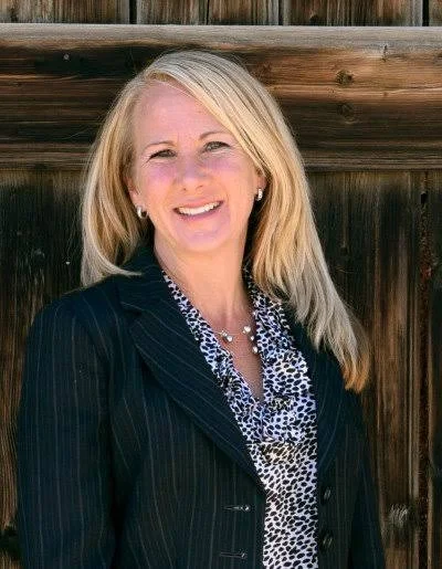 A woman with blonde hair smiling, wearing a dark pinstripe blazer and patterned blouse, standing outside in front of a wooden fence.